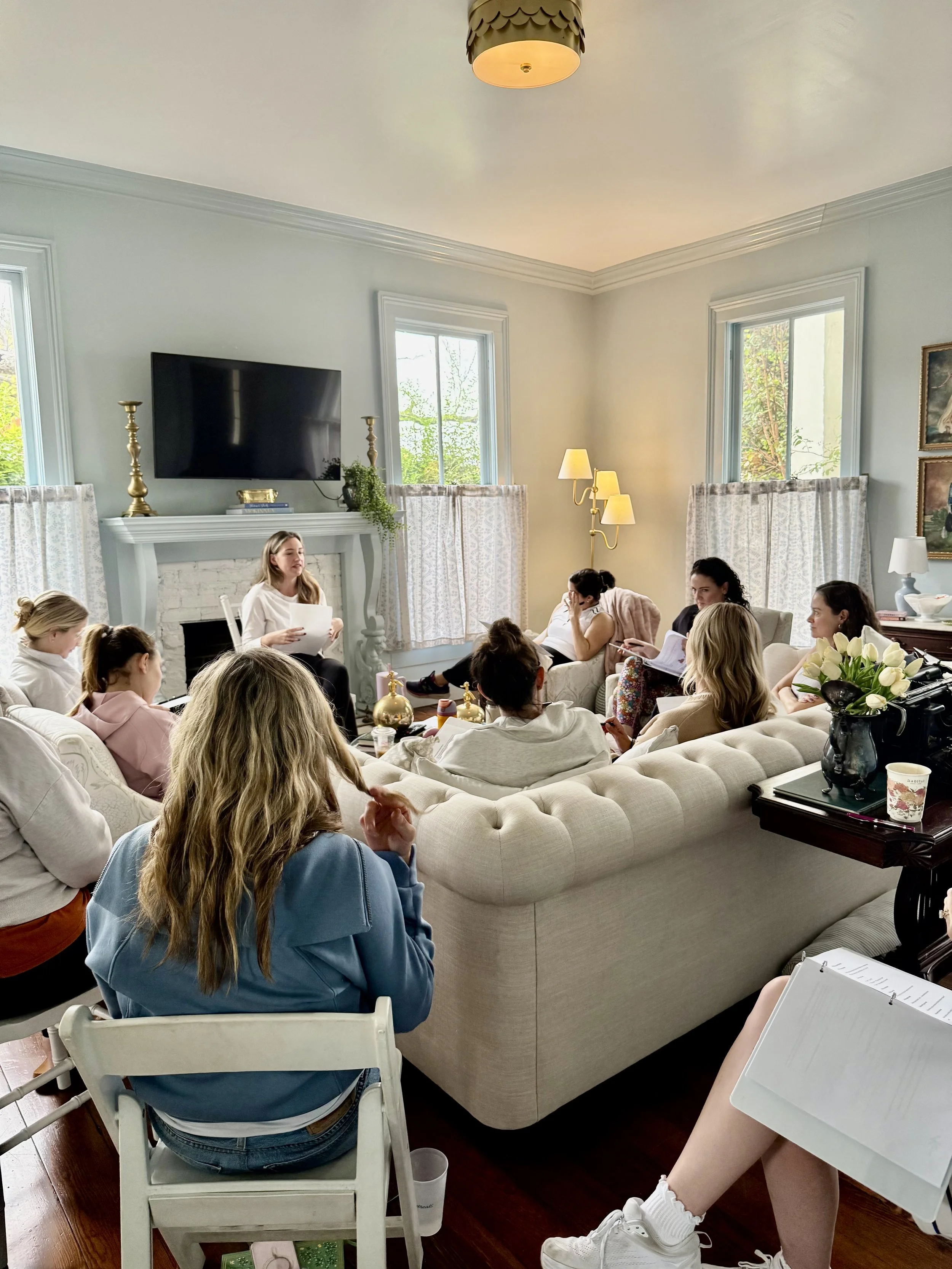 A group of women and girls attending a meeting or class in a cozy living room with white walls, large windows, and a fireplace. One woman is standing by the fireplace, speaking to the group.