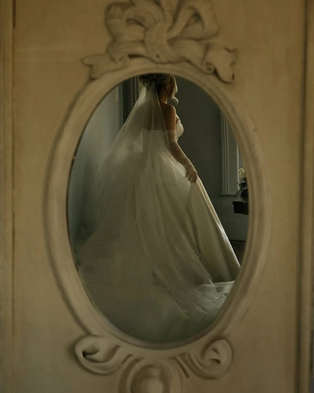 A bride in a wedding gown with a veil stands near a window, seen through an ornate oval mirror at The Bingham Estate luxury hotel outside of Dallas, TX. This luxury mckinney hotel is the perfect place for photography, bridal showers, group events.