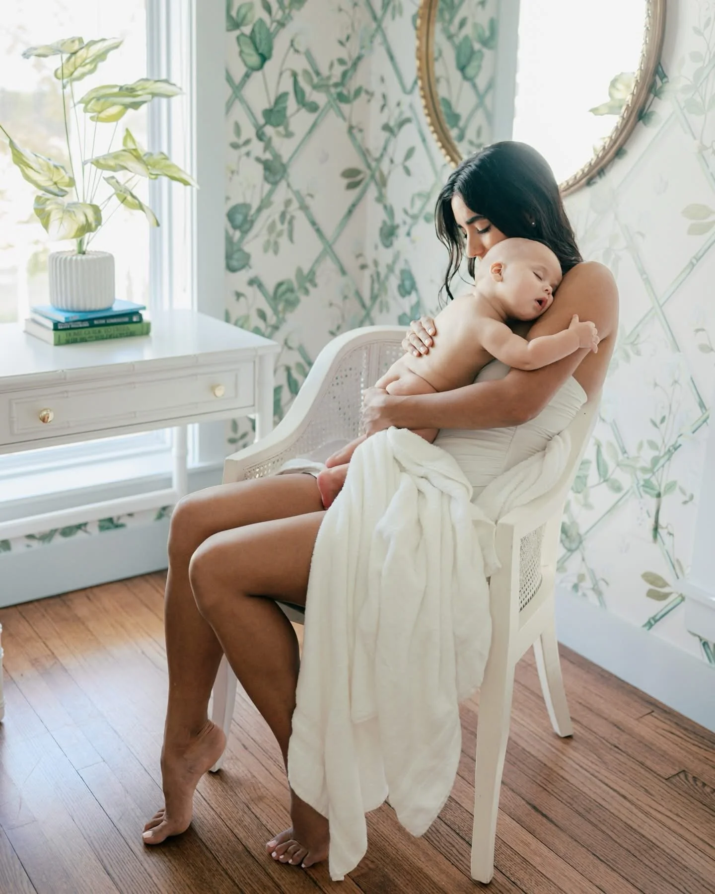 A woman in a white dress sitting on a chair, breastfeeding a sleeping baby in a bright room with floral wallpaper, wooden floor, and a white table with a plant and books.