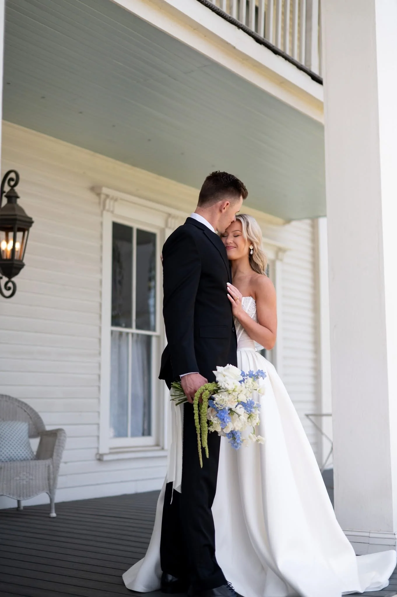 A bride and groom embrace on a porch, with the bride holding a bouquet of white and blue flowers, and the groom wearing a black suit.