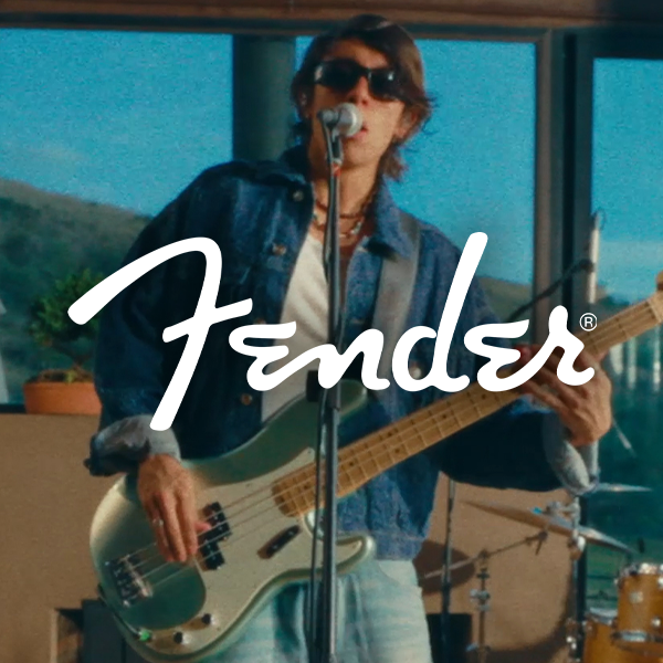 A person with shoulder-length hair wearing sunglasses, a denim jacket, and white pants, playing a bass guitar and singing into a microphone in a studio with a scenic window view.