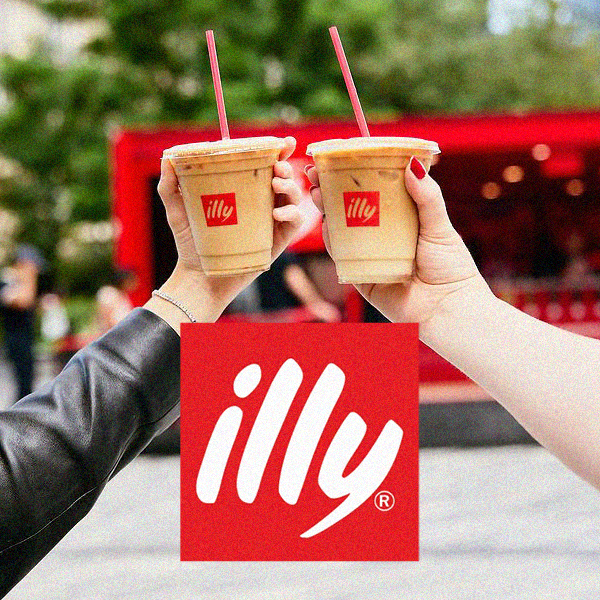 Two people holding iced coffee drinks with straws, raised in a toast, with an illy coffee logo displayed in front of a red truck and green trees in the background.