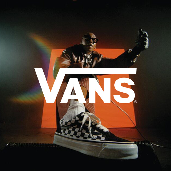 A person wearing checkered Vans sneakers sitting on a platform with the Vans logo displayed in front of them, set against a dark background with a colorful rainbow light effect.