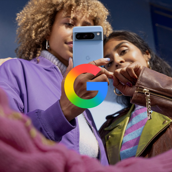 Two young women taking a selfie with a Google smartphone. One woman has curly blonde hair and is wearing a purple jacket, while the other has dark hair and is wearing a green jacket with a colorful striped shirt. They are outdoors and smiling.