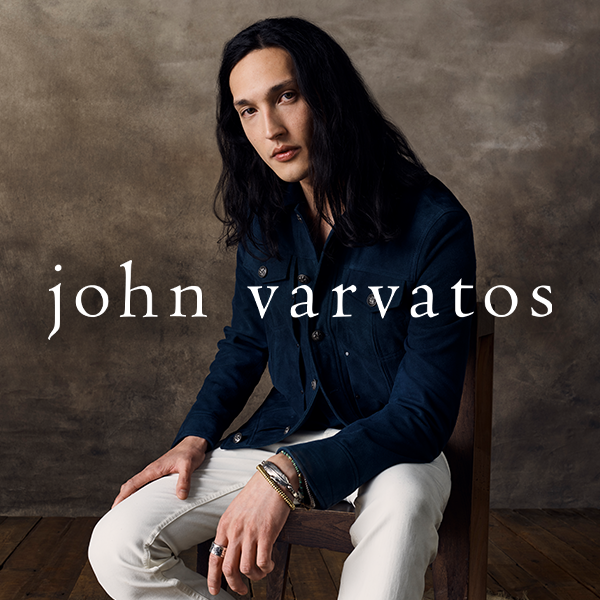 A woman with long dark hair sitting on a chair, wearing a navy blue jacket and white pants, against a plain brown background with the text "john varvatos" across her chest.