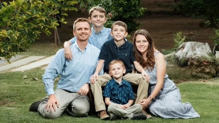 A family of six posing outdoors at a tropical location with greenery and a thatched roof structure in the background, smiling at the camera.