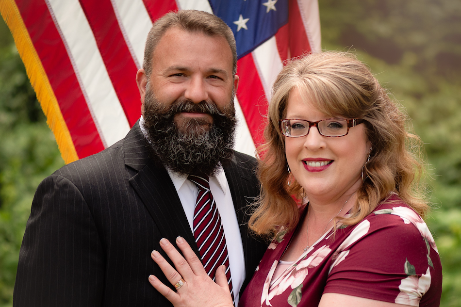 A man and woman standing together outdoors with the American flag in the background, smiling at the camera.