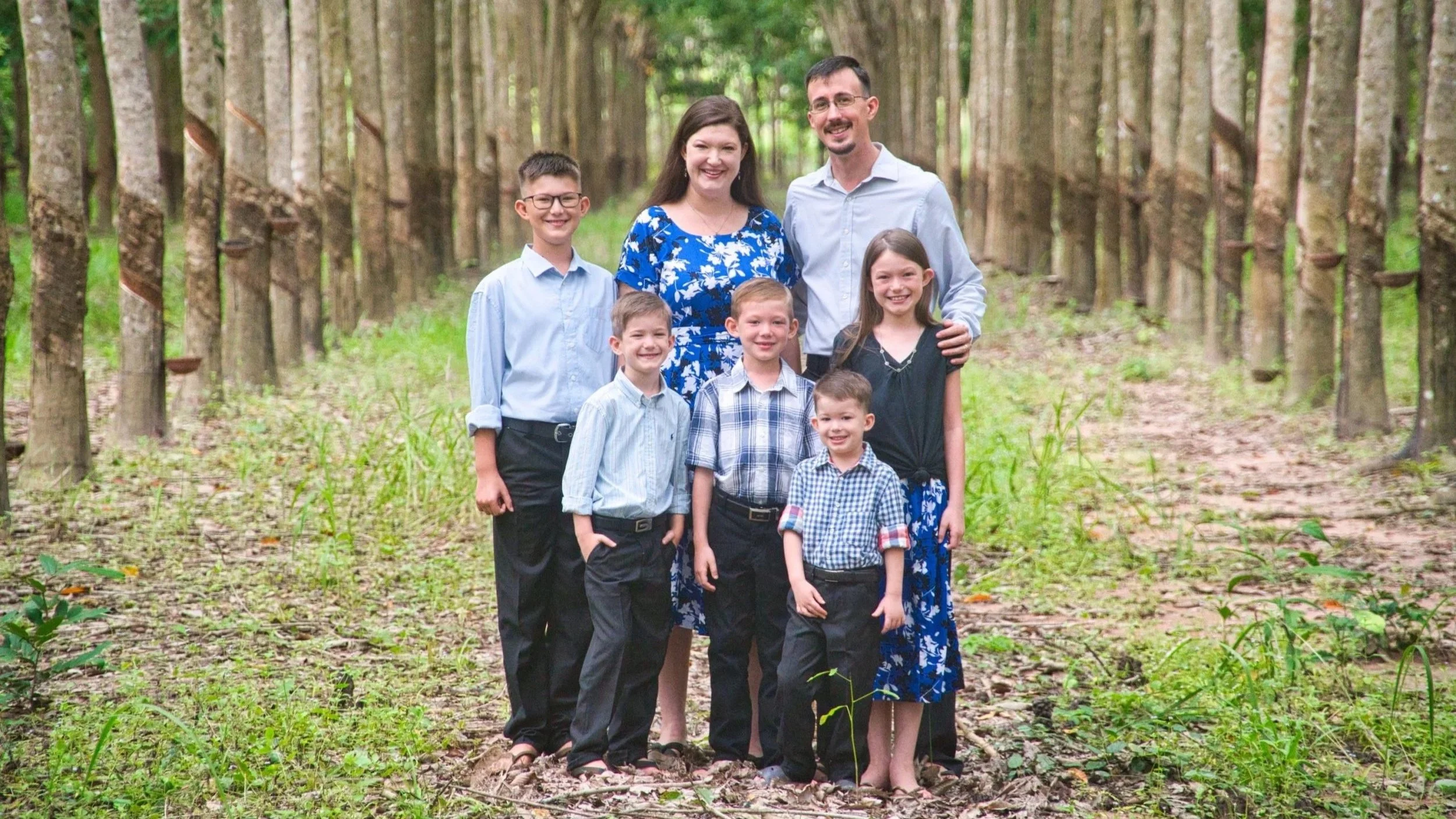 A happy family of seven outdoor in a park with trees in the background, including two adults and five children, posing for a photo.