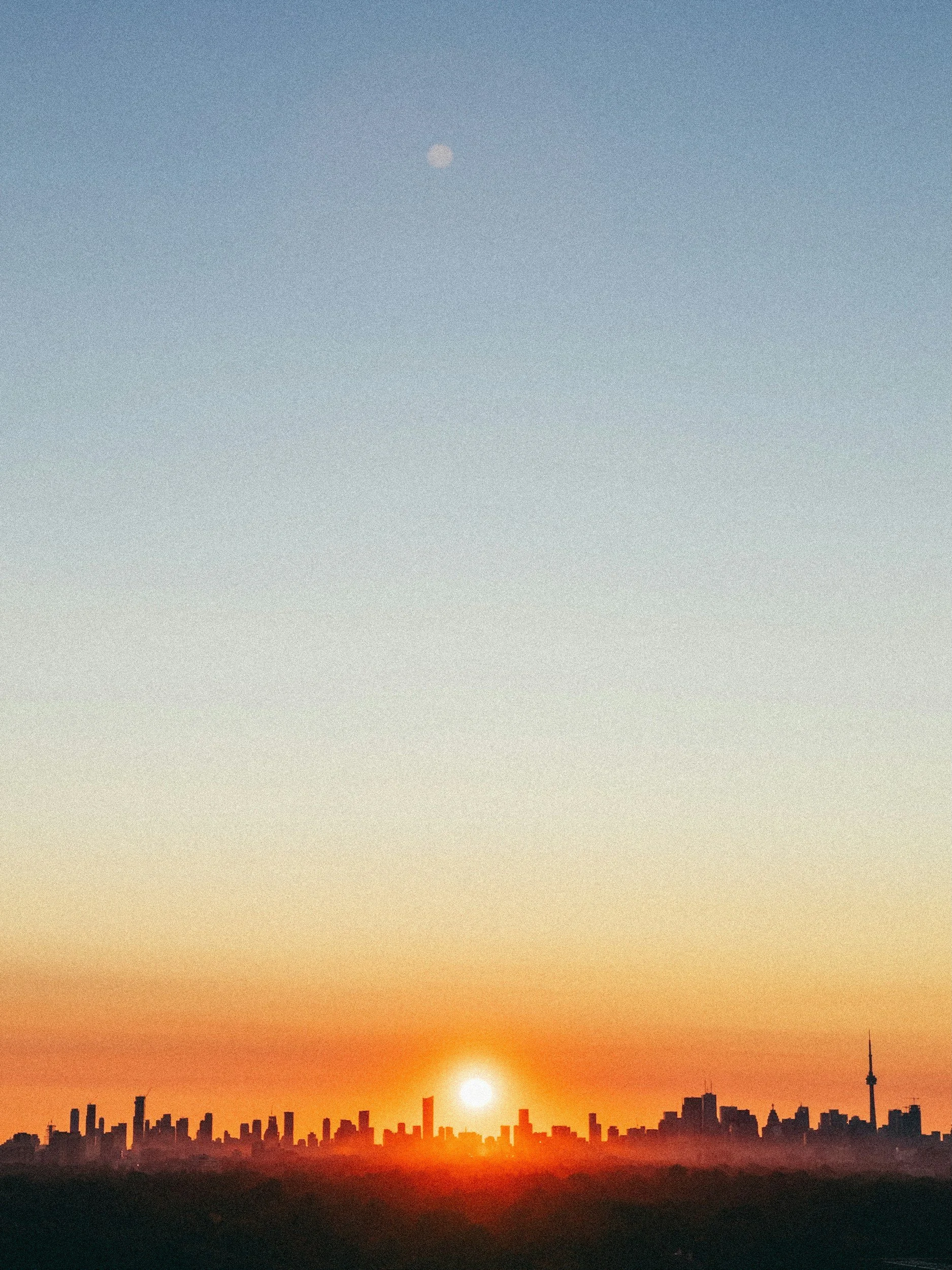 Sun setting over a city skyline at dusk with a visible moon in the sky.