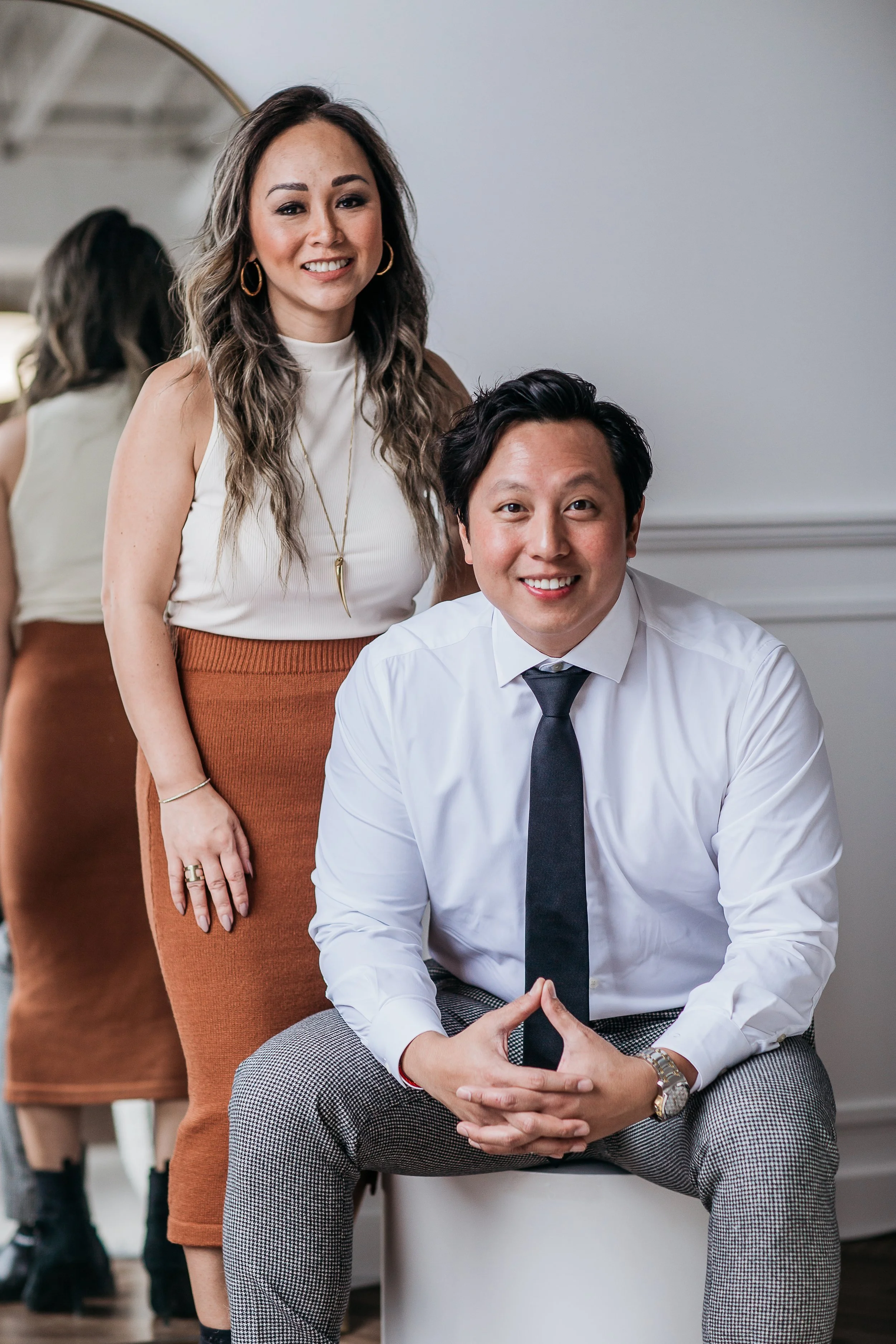 Two people, a woman and a man, posing indoors for a photo. The woman is standing with her hand resting on the shoulder of the man, who is sitting on a stool. The woman has long wavy hair, wears a white sleeveless top, orange-brown skirt, and gold jewelry. The man has short dark hair, wears a white shirt, gray checkered pants, and a black tie, with a watch on his left wrist. Both are smiling at the camera.