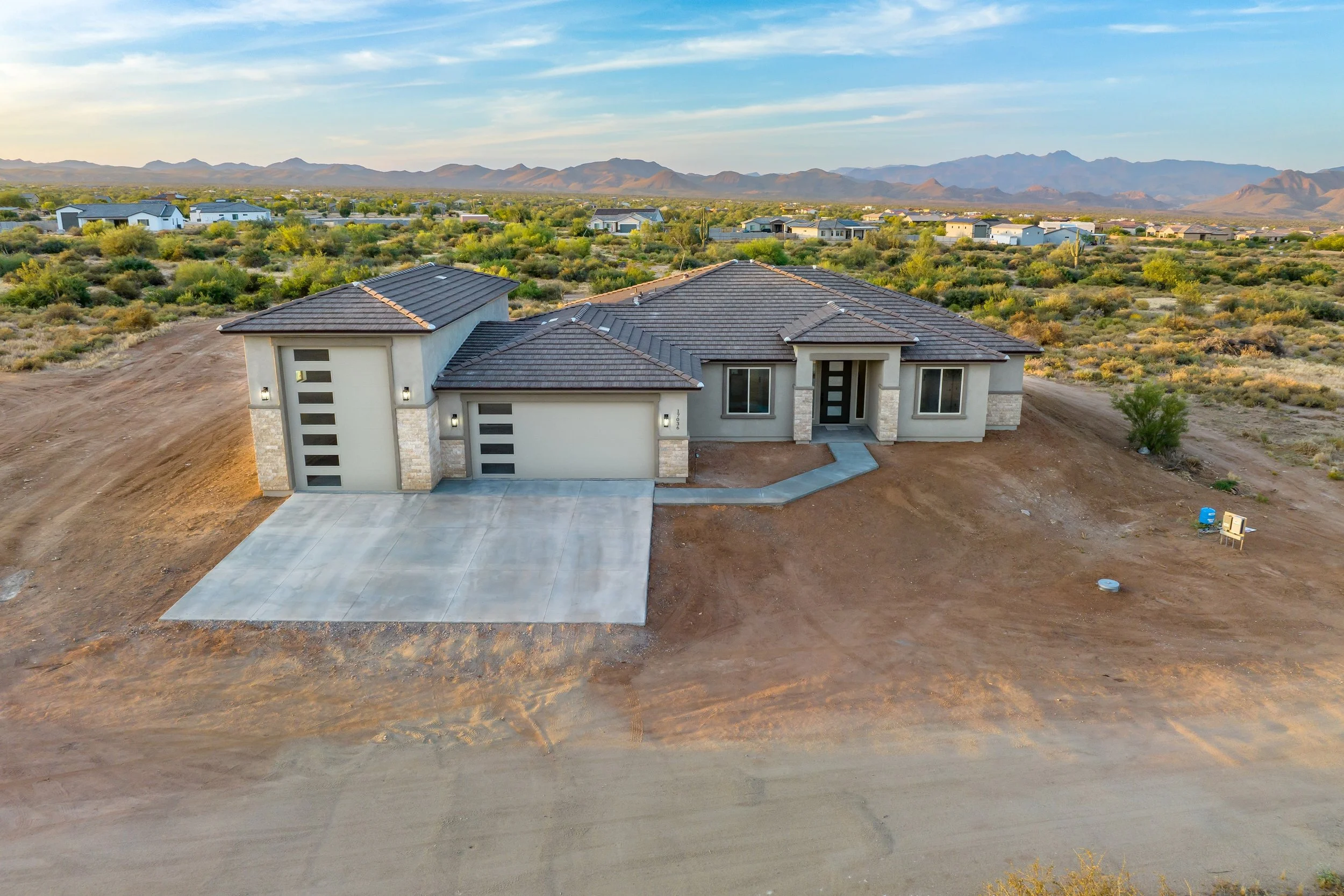A newly constructed single-story house with a gray tiled roof, white exterior walls, and a concrete driveway, situated in a desert landscape with mountains in the background.