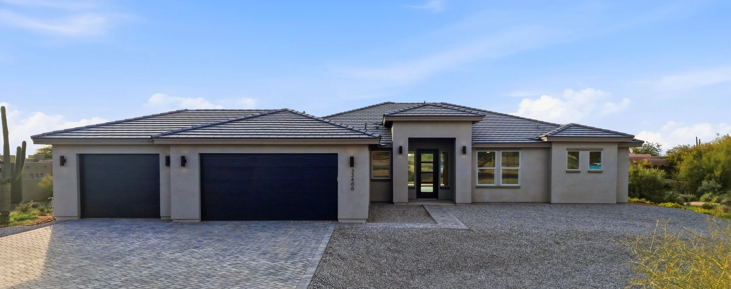 Modern single-story house with a gray tiled roof, white exterior walls, black garage doors, and black-framed glass front door, surrounded by desert landscaping.