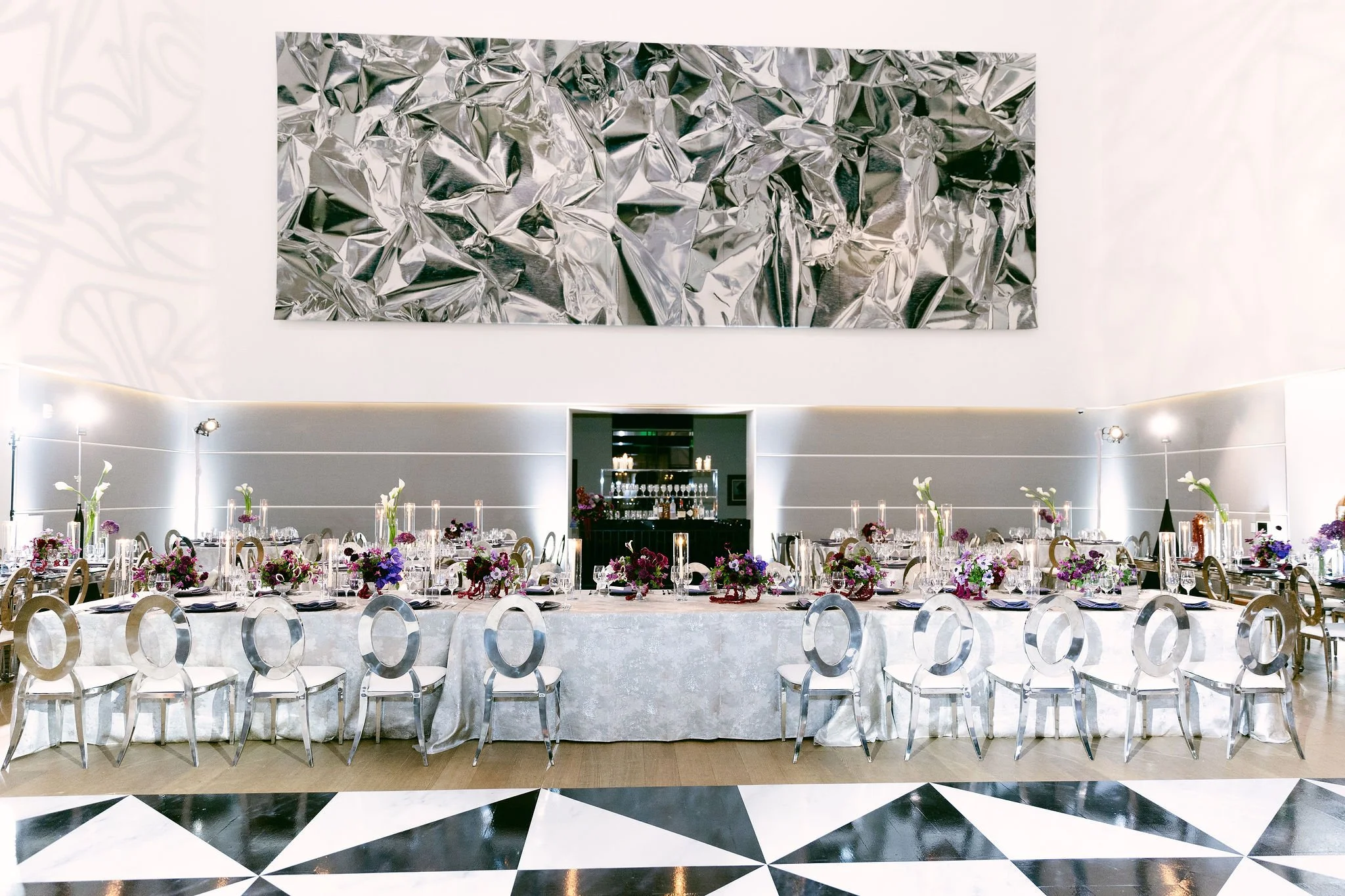 Elegant banquet hall with a long table adorned with floral centerpieces and silver chairs, set for a formal event, with a modern abstract metal art piece on the wall