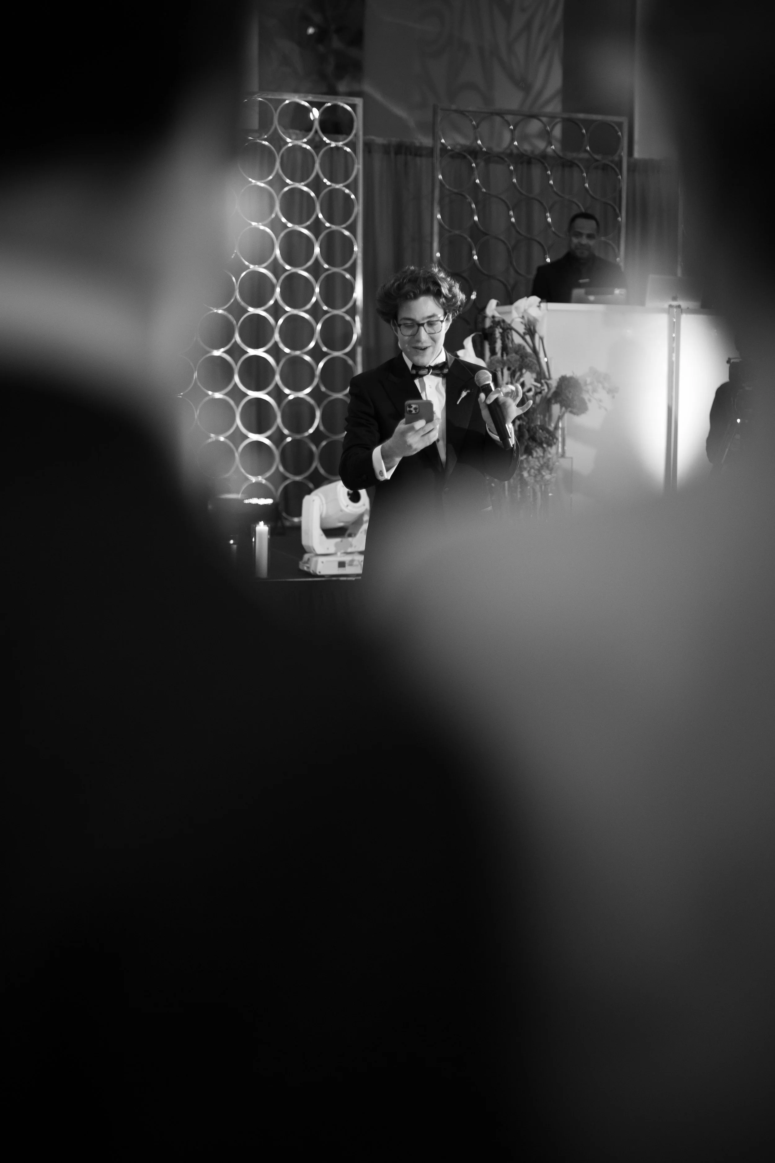 A person in a tuxedo with glasses is standing on a stage, speaking into a microphone and looking at a phone, with a DJ in the background.