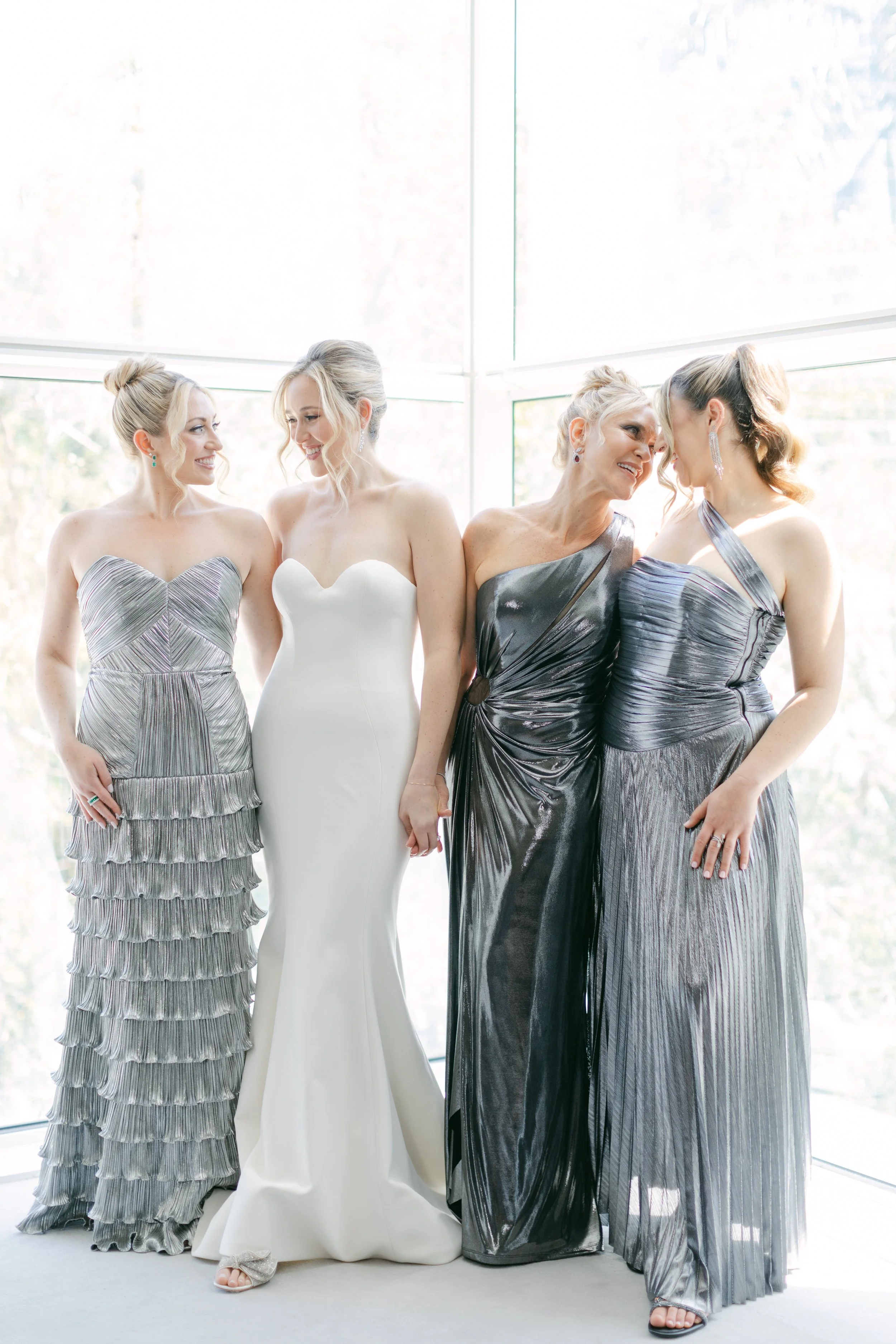 Four women in formal dresses standing together in front of a large window with natural light, smiling and interacting with each other.