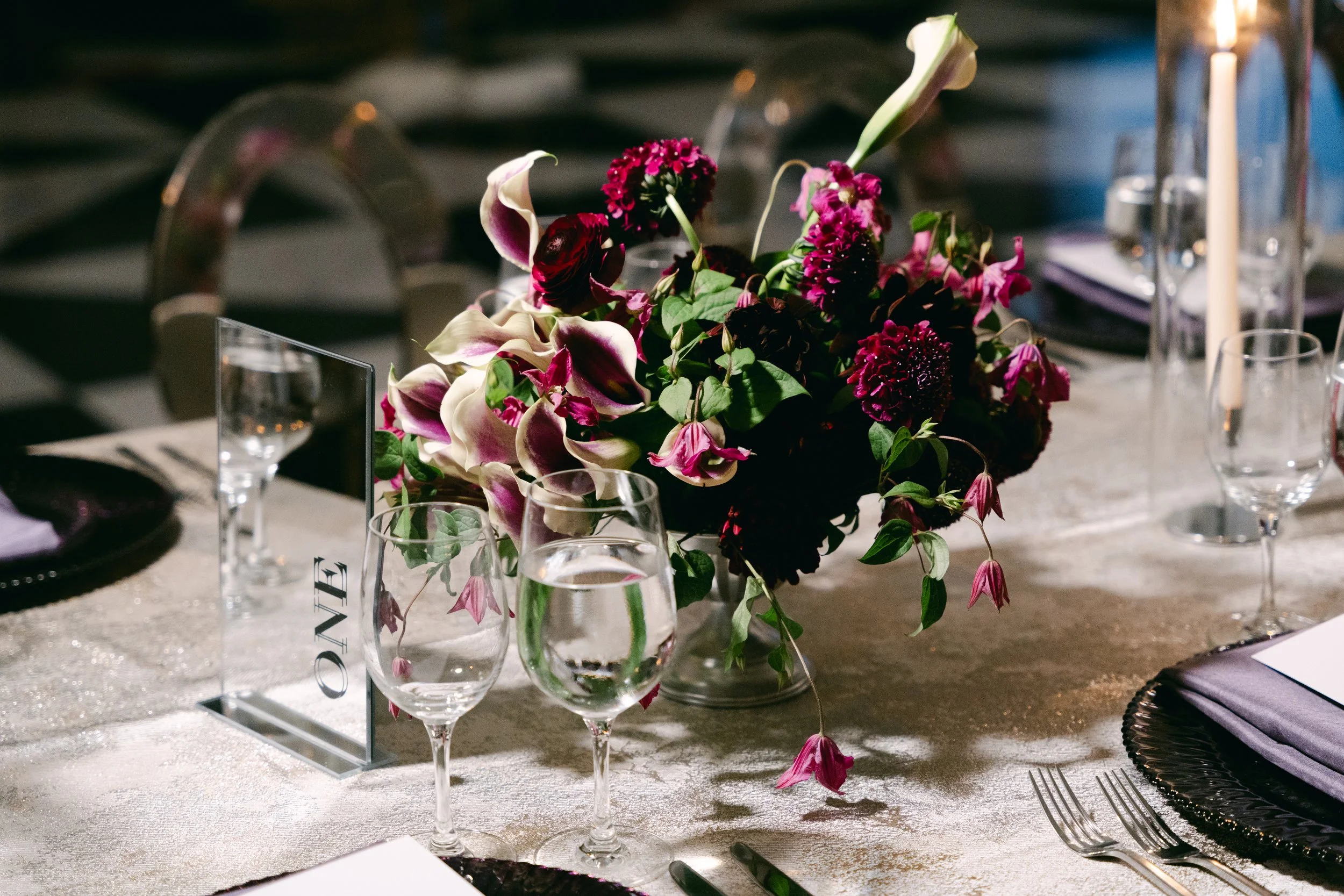 A beautifully decorated banquet table with a central flower arrangement of purple, pink, and white flowers, set with wine glasses, water glasses, plates, forks, and napkins, with a white tablecloth and candles.