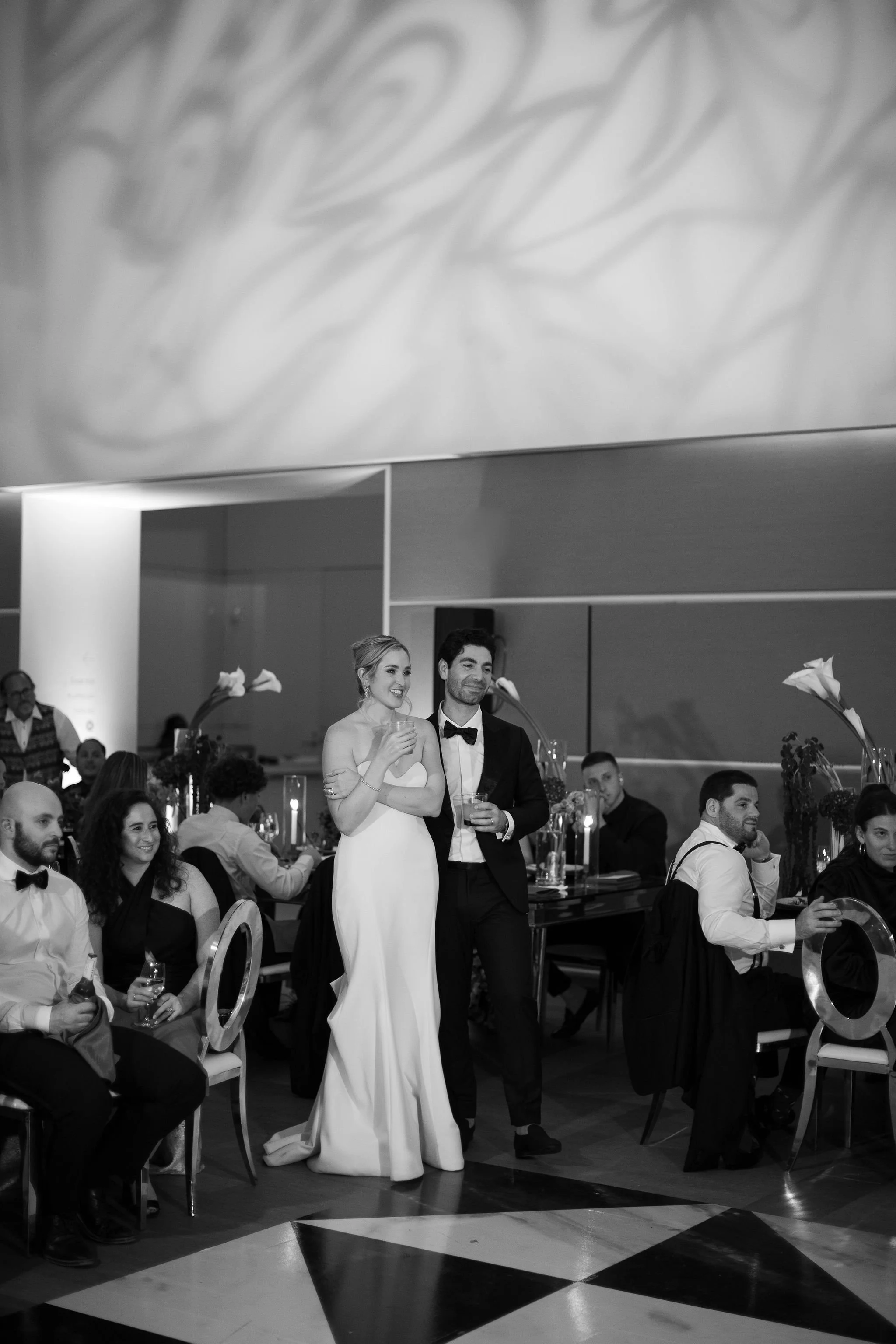Black and white photo of a wedding reception with a bride and groom standing together and smiling, surrounded by guests seated at tables with floral centerpieces and candles.