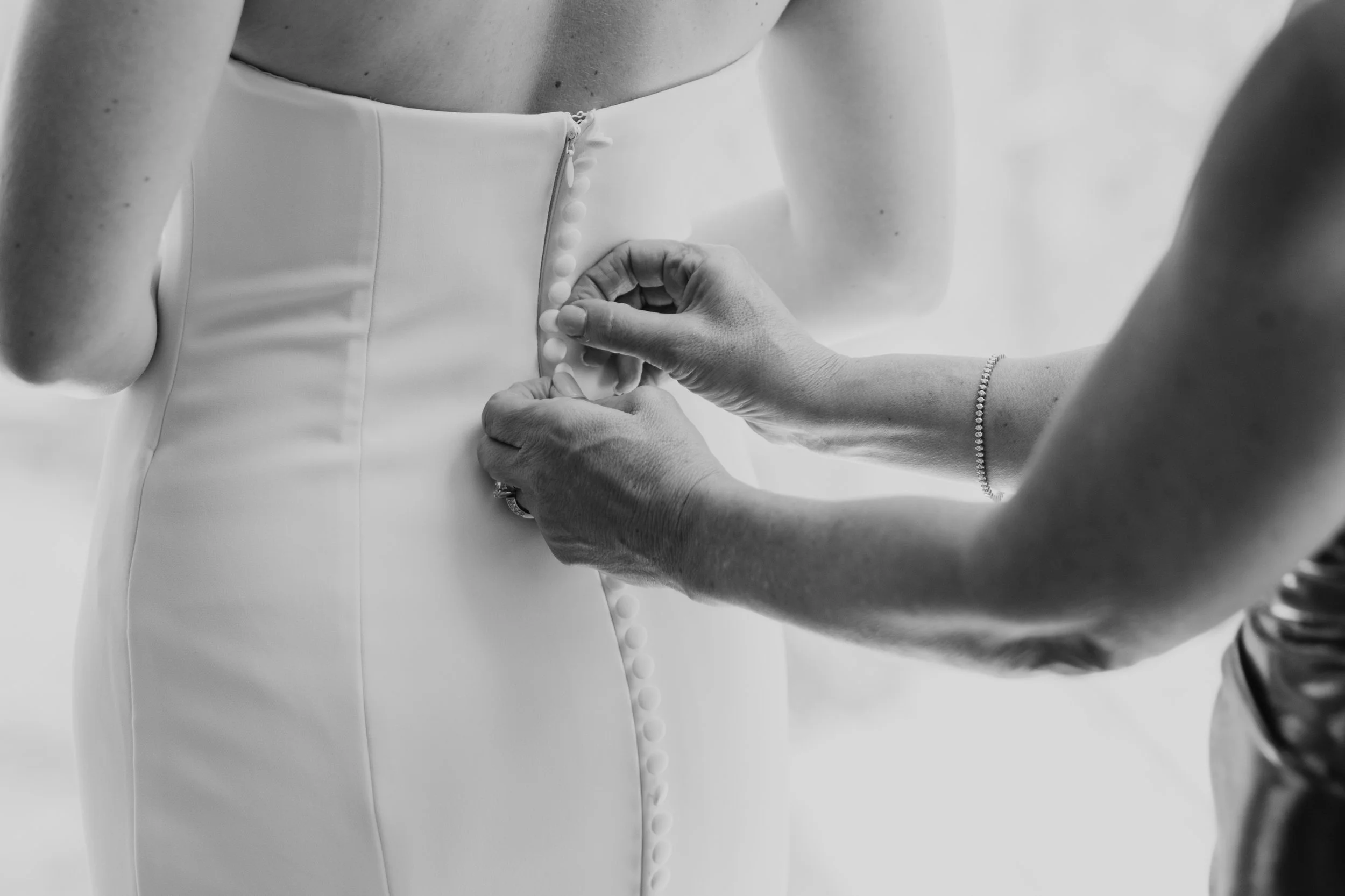A person is fastening buttons on the back of a wedding dress, which is being worn by another person.