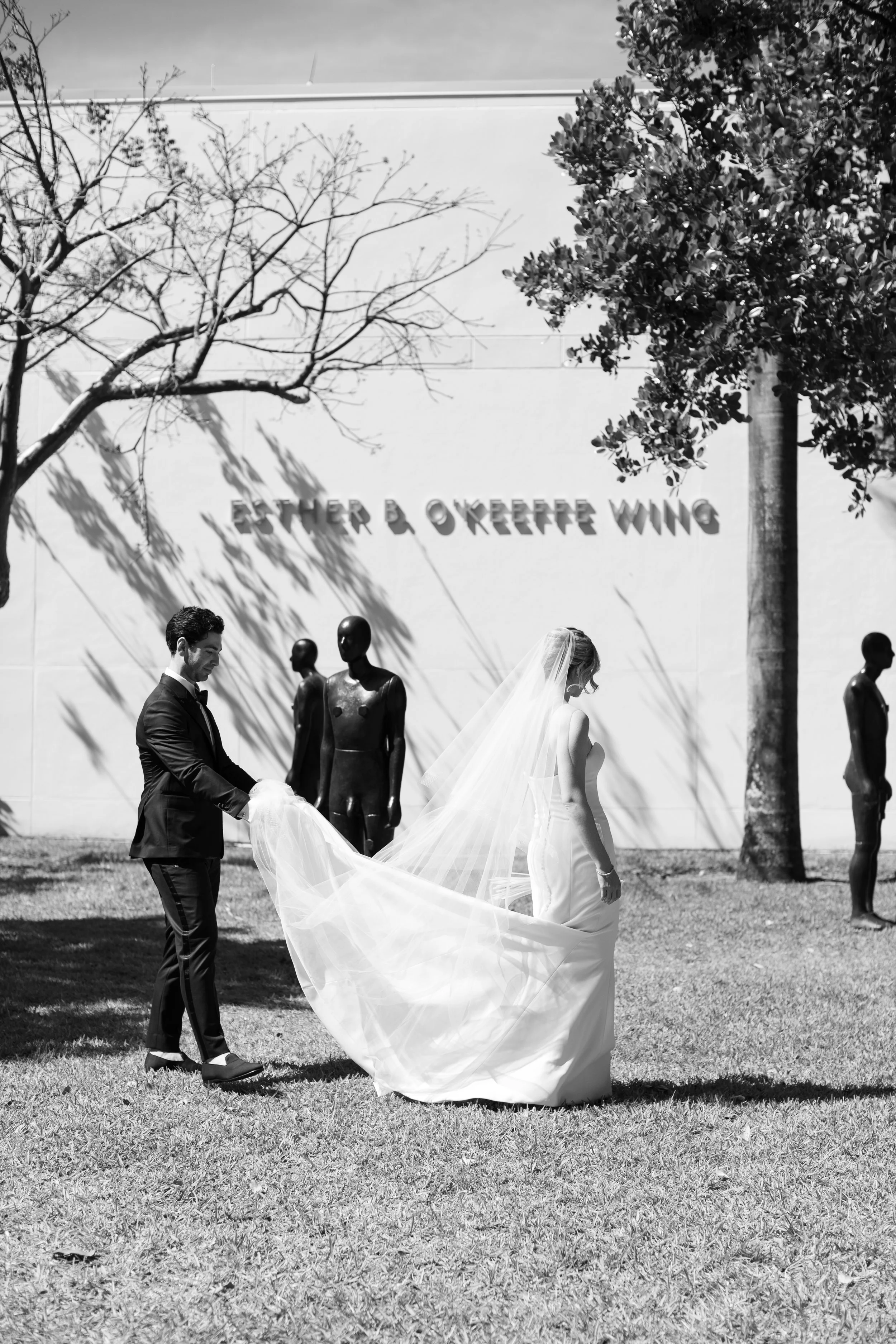 A bride and groom in wedding attire, outdoors on grass, with trees and sculptures in the background. The groom is holding the bride's train.
