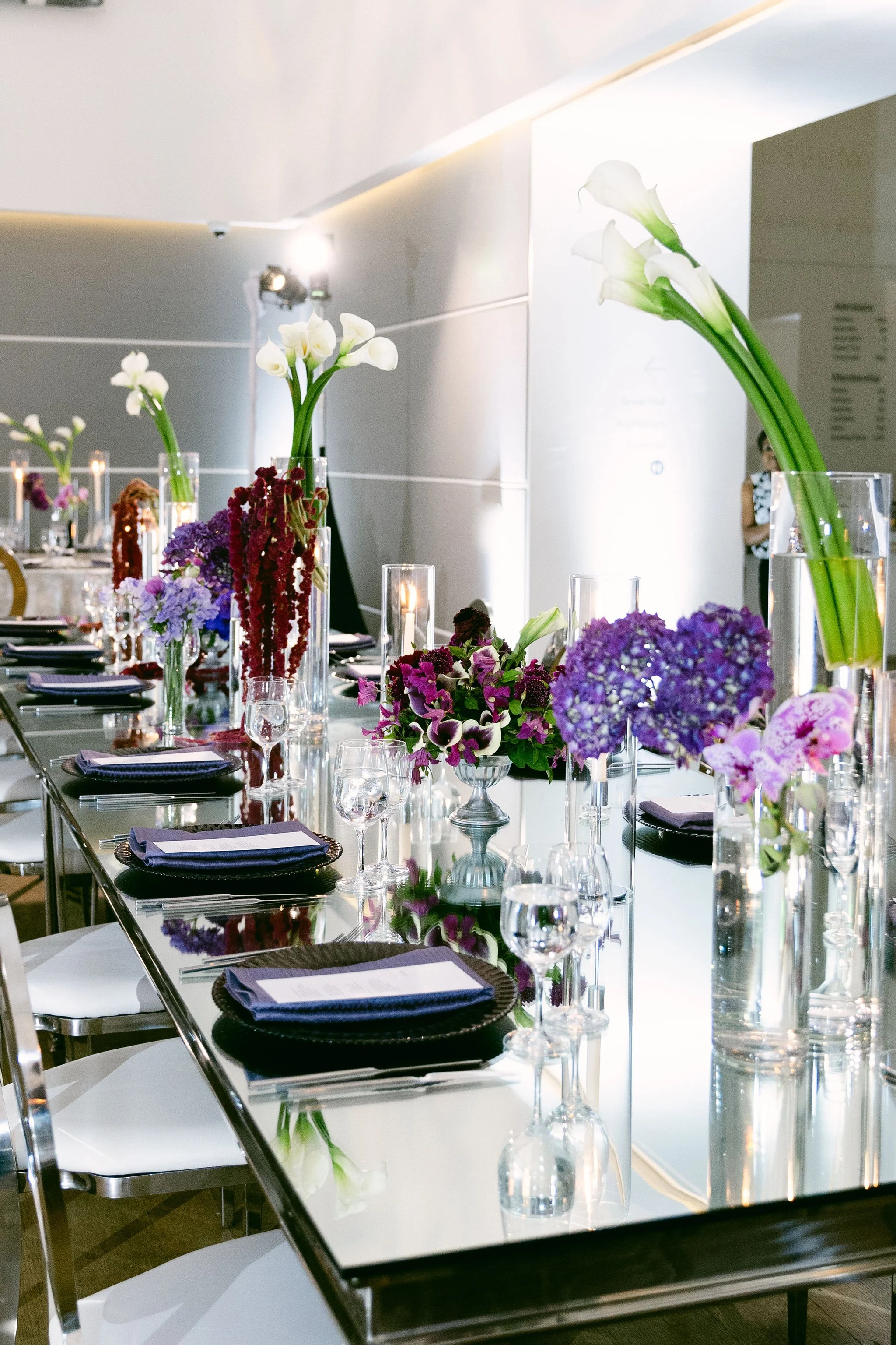 Elegant dining table set with tall vases of white calla lilies, purple and pink hydrangeas, and other colorful flowers, with black napkins on white plates, all arranged in a modern indoor setting.