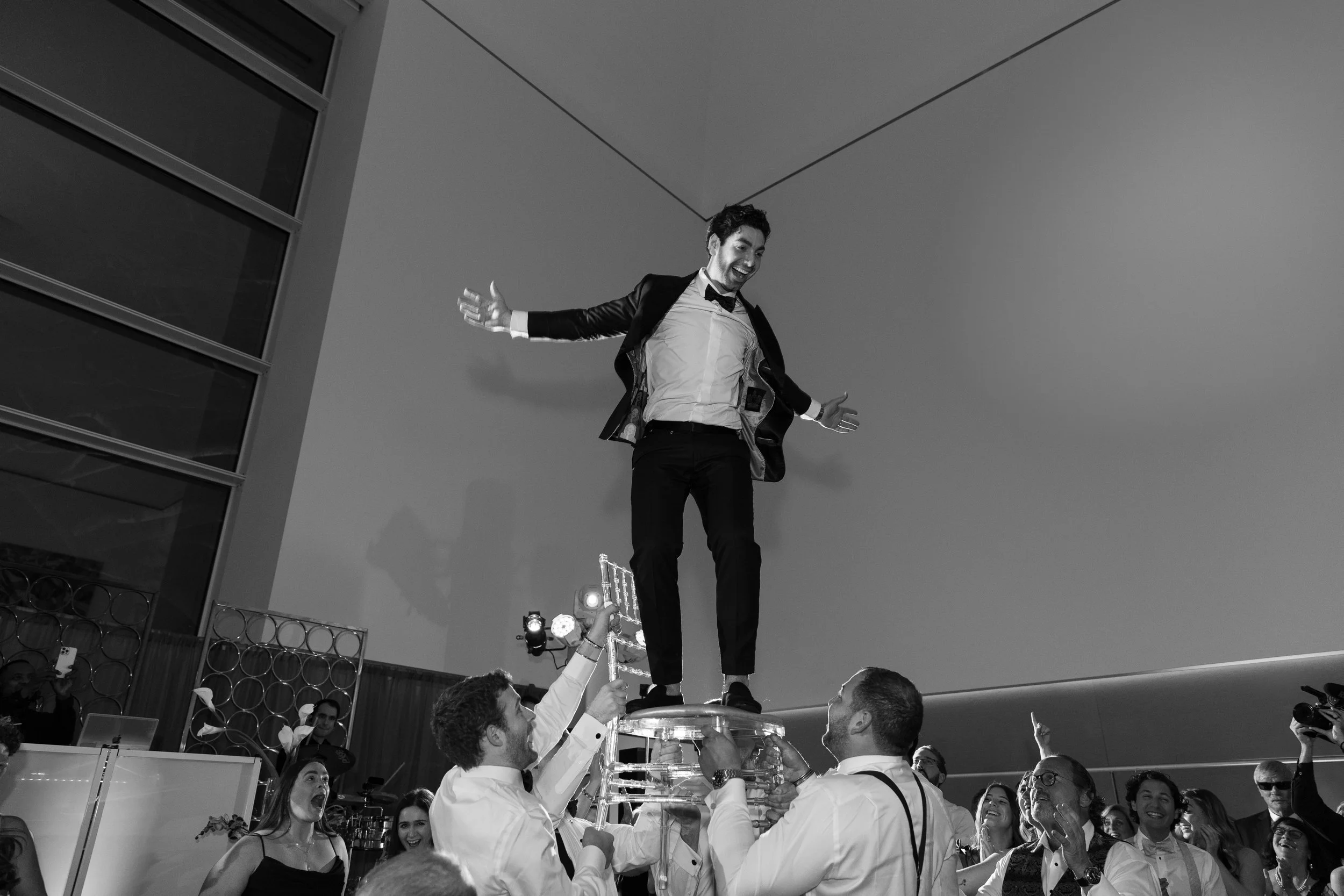 A man in a tuxedo stands on a glass platform held by several people at an event, smiling and gesturing with arms outstretched, surrounded by a crowd.