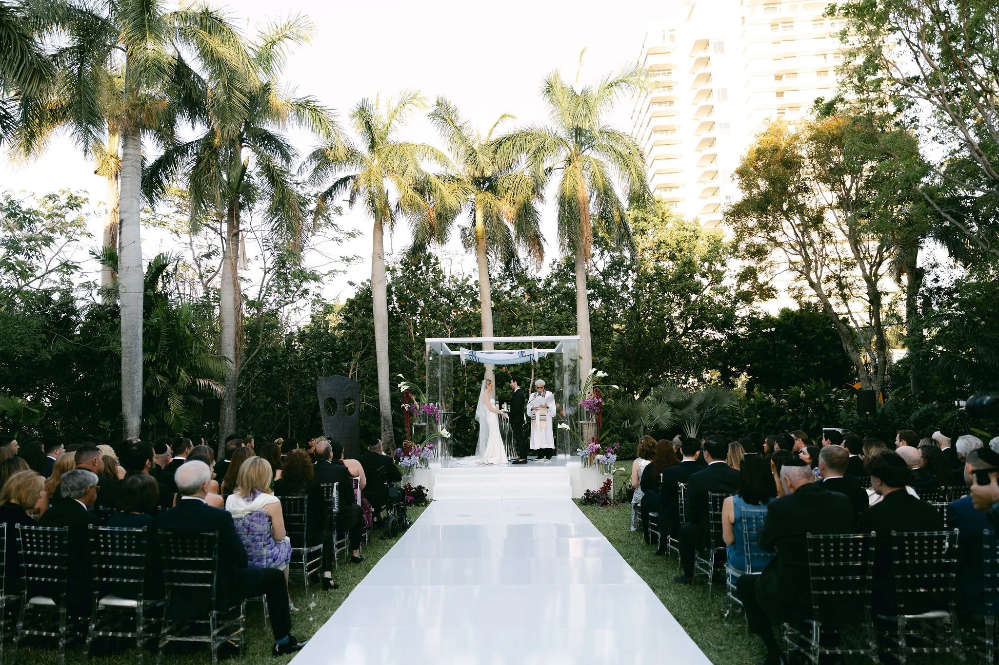 Outdoor wedding ceremony with a bride, groom, and officiant under a canopy, surrounded by tall palm trees and lush greenery, with guests seated on either side of a white aisle in a city setting with high-rise buildings in the background.