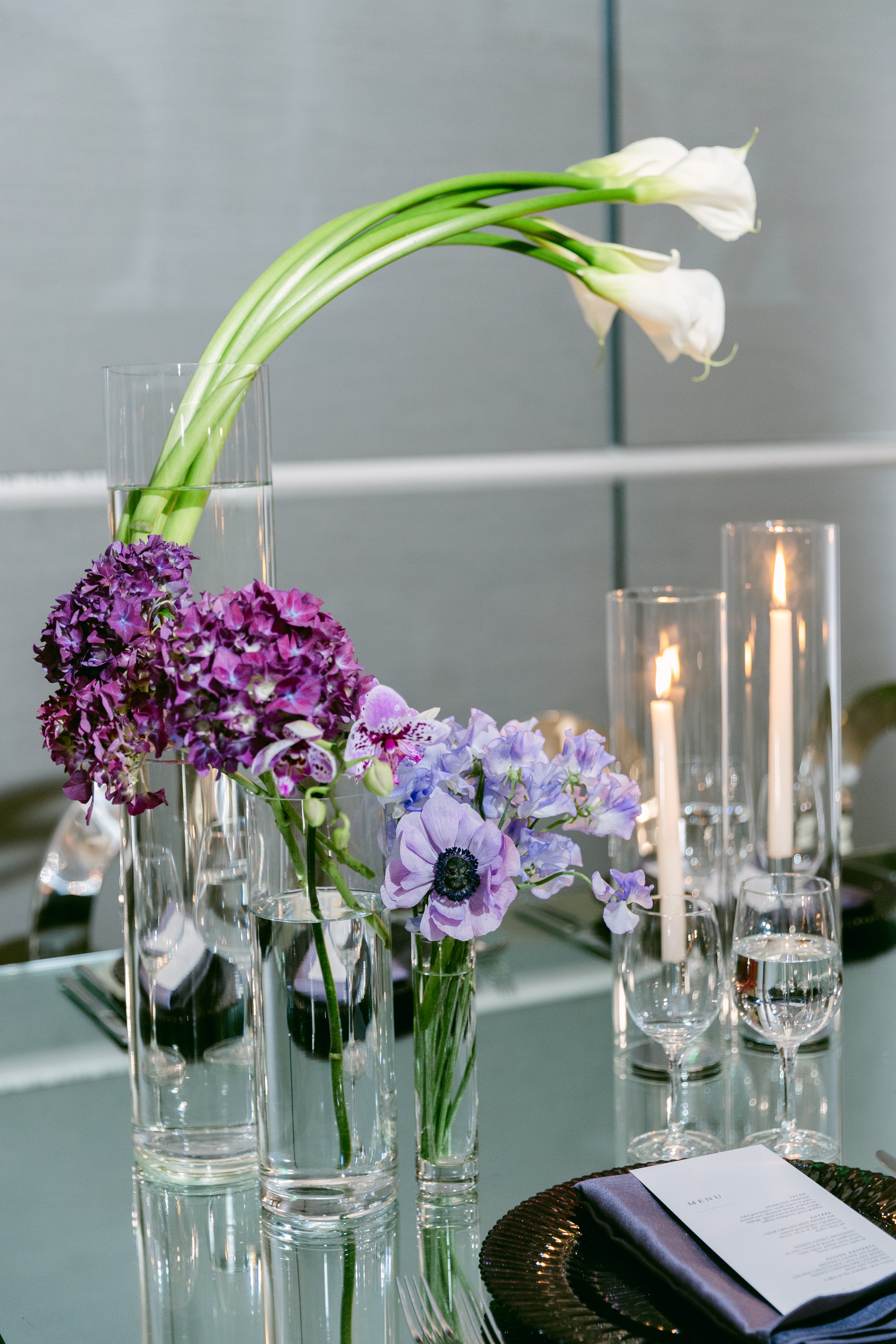 Elegant table setting with tall glass vases holding white calla lilies, purple hydrangeas, and light purple anemones, with lit candles in glass holders, a menu, and glassware.