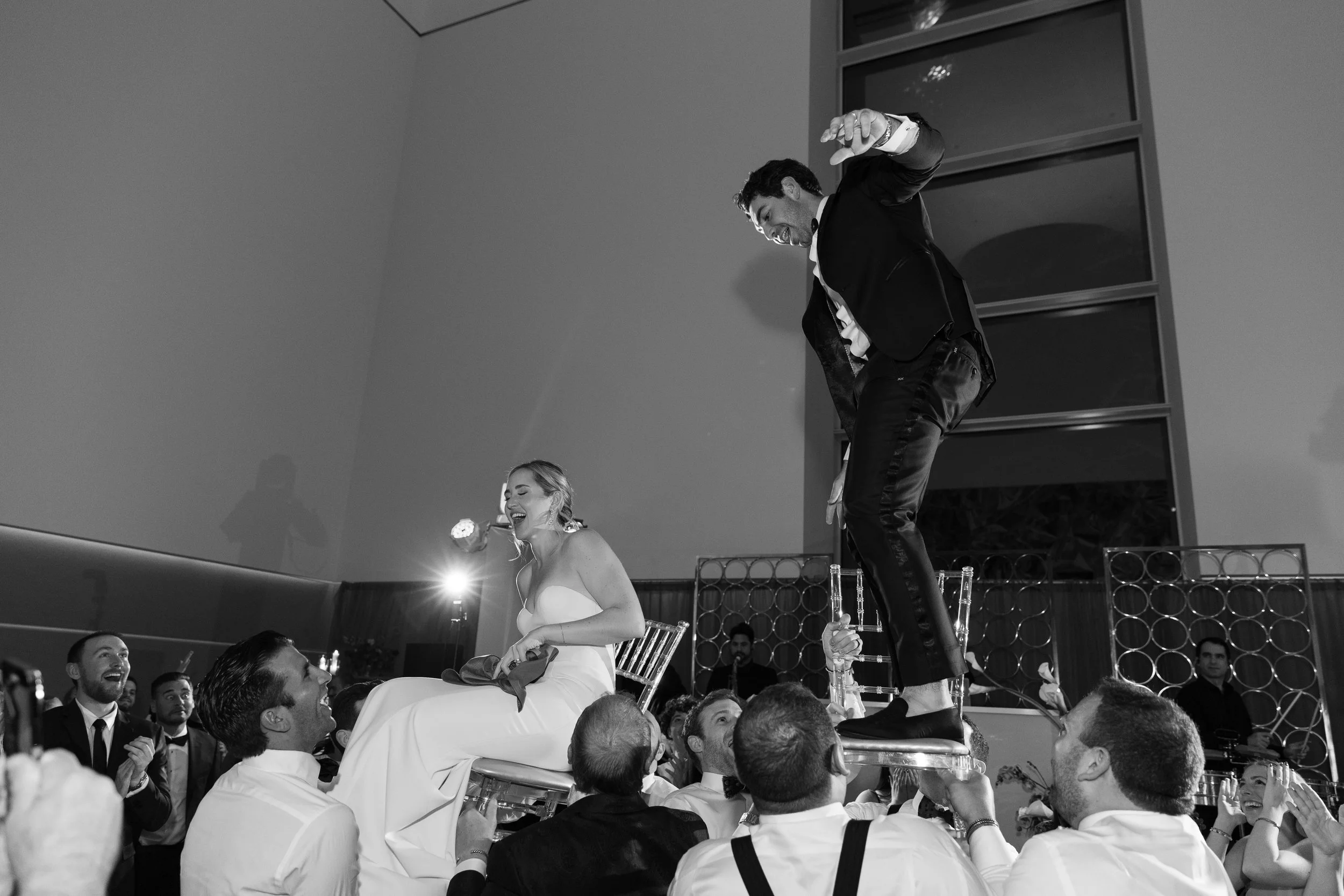 A wedding reception with guests lifting the bride and groom on chairs during a celebration, with the bride and groom smiling and wedding guests clapping and cheering.