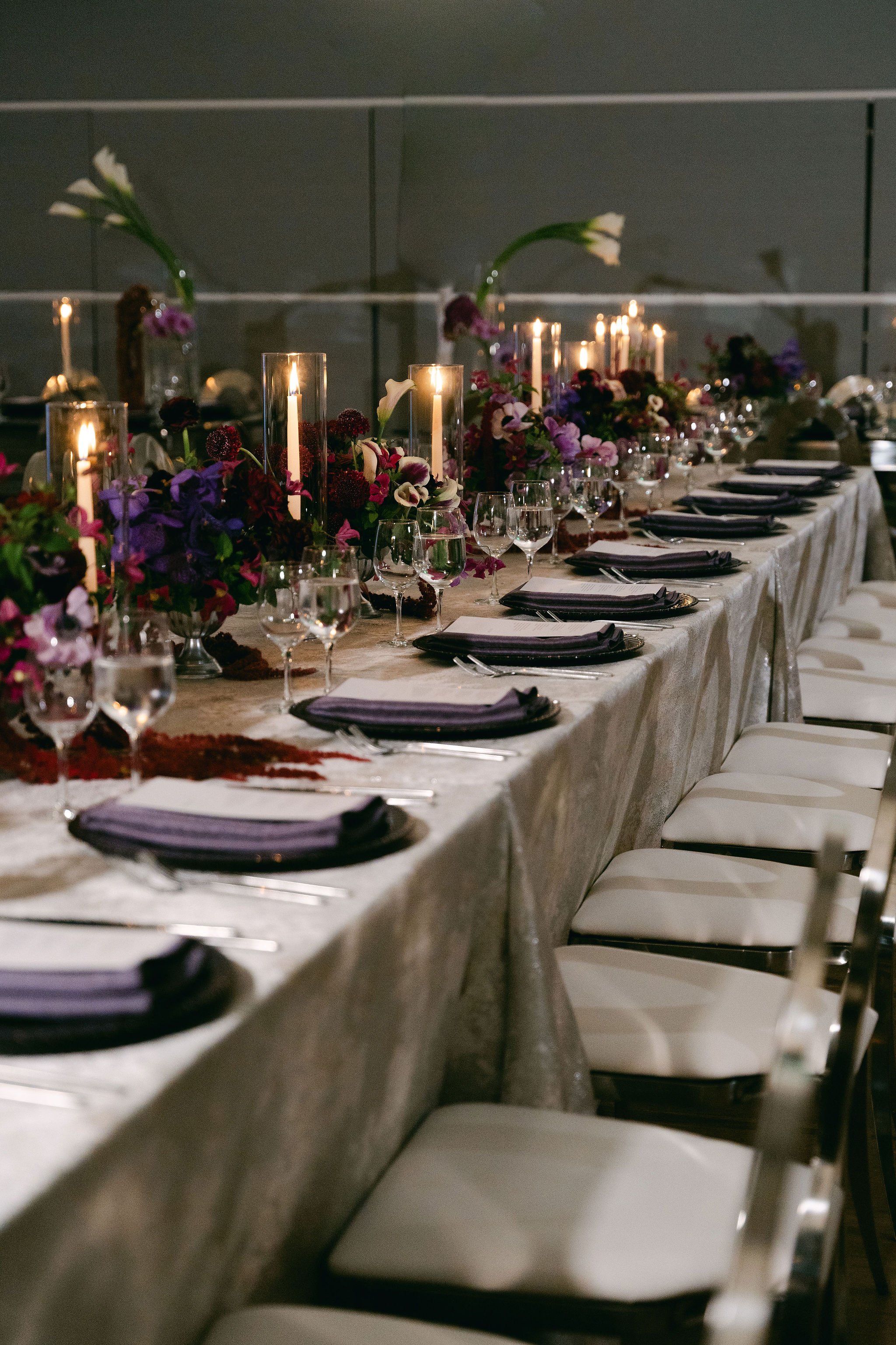 Decorated long dining table with floral centerpieces, candles, wine glasses, and black napkins set for an event or wedding reception.