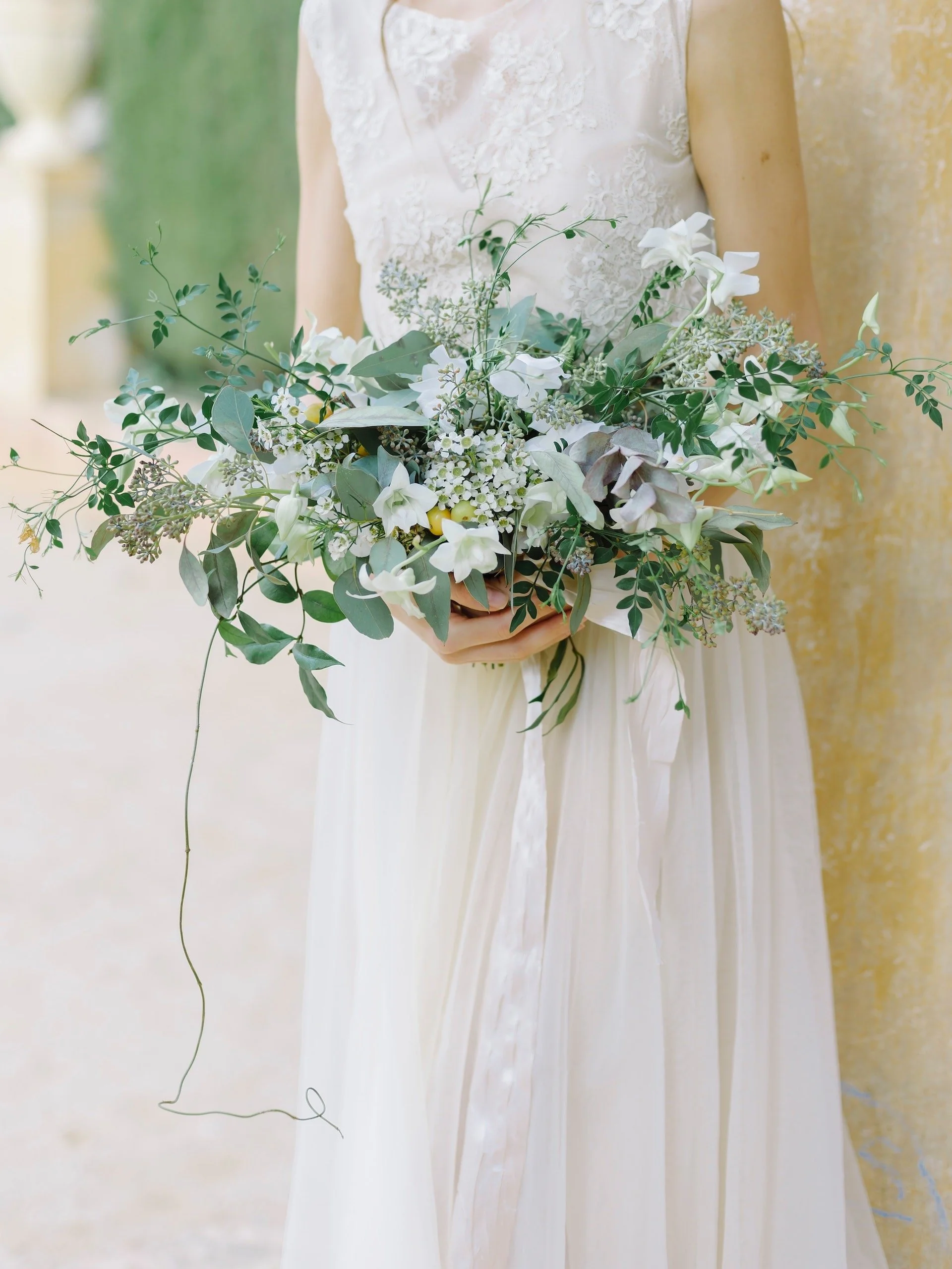 A woman in a white lace wedding dress holding a bouquet of white and green flowers and foliage, standing outdoors.