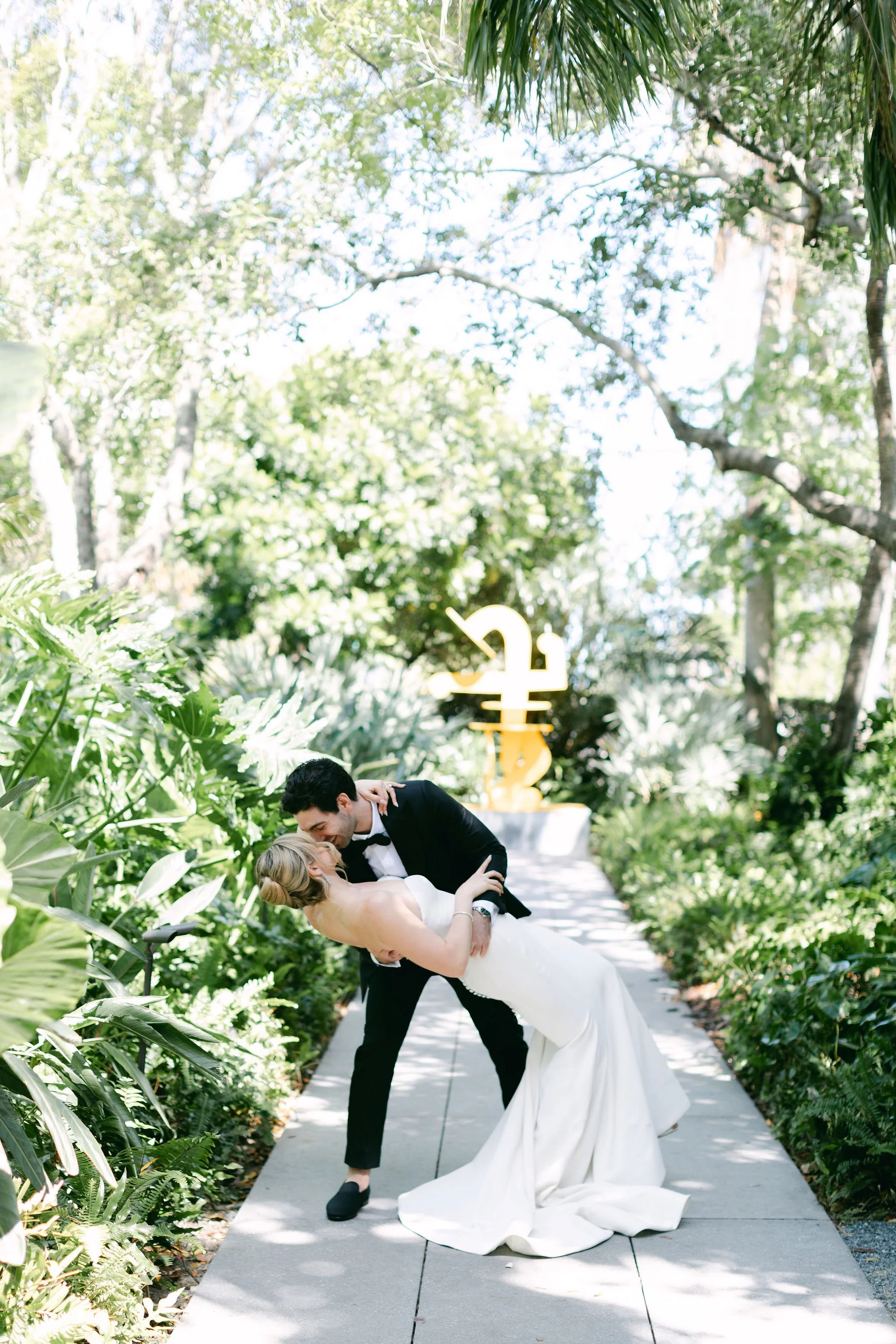 A bride and groom sharing a dance on a garden pathway during their wedding, with lush greenery and a yellow abstract sculpture in the background.