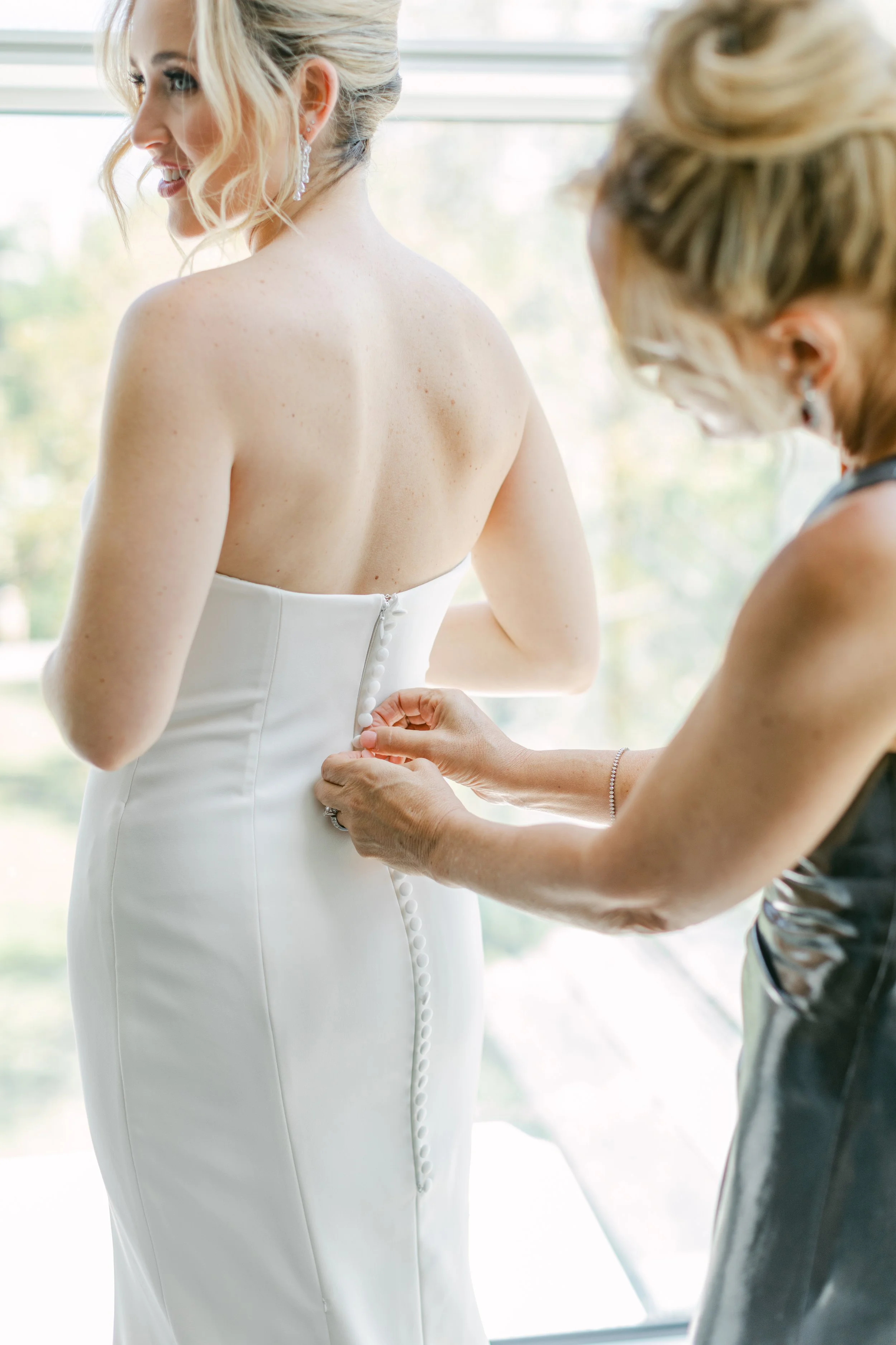 A bride is being assisted in buttoning her wedding dress, standing in front of a window with natural light.
