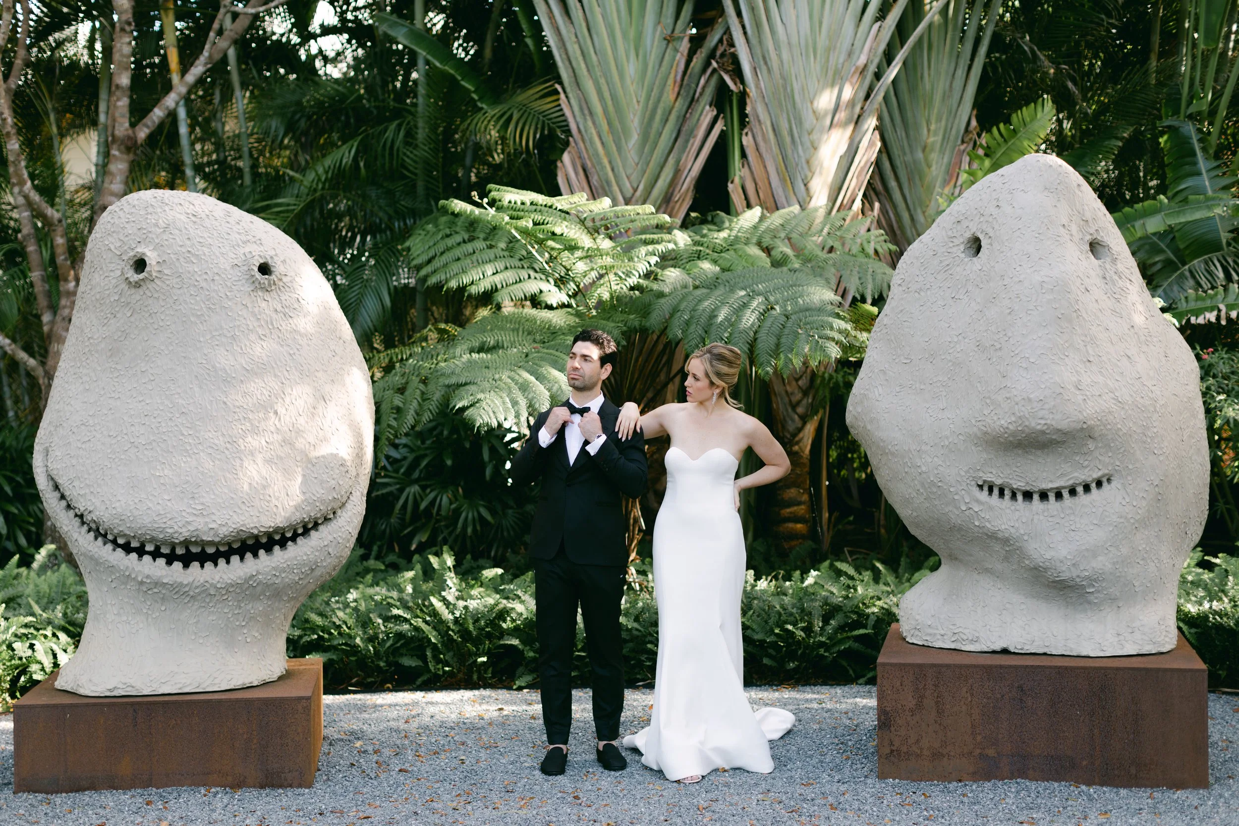 A man in a black tuxedo and a woman in a white strapless wedding dress stand between two large abstract sculptures of smiling faces in a lush, green garden setting.