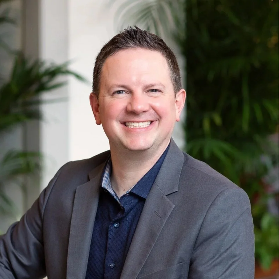 Dean is smiling, wearing a gray blazer, in front of a slightly blurred background with greenery.