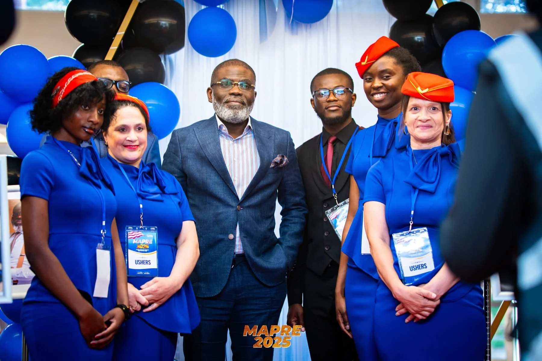 Six people standing together at a conference, with balloons and a « MAPRE 2025 » sign in the background. Four women wearing matching blue dresses and red headbands, two men in formal attire. The women have badges labeled « Ushers ».
