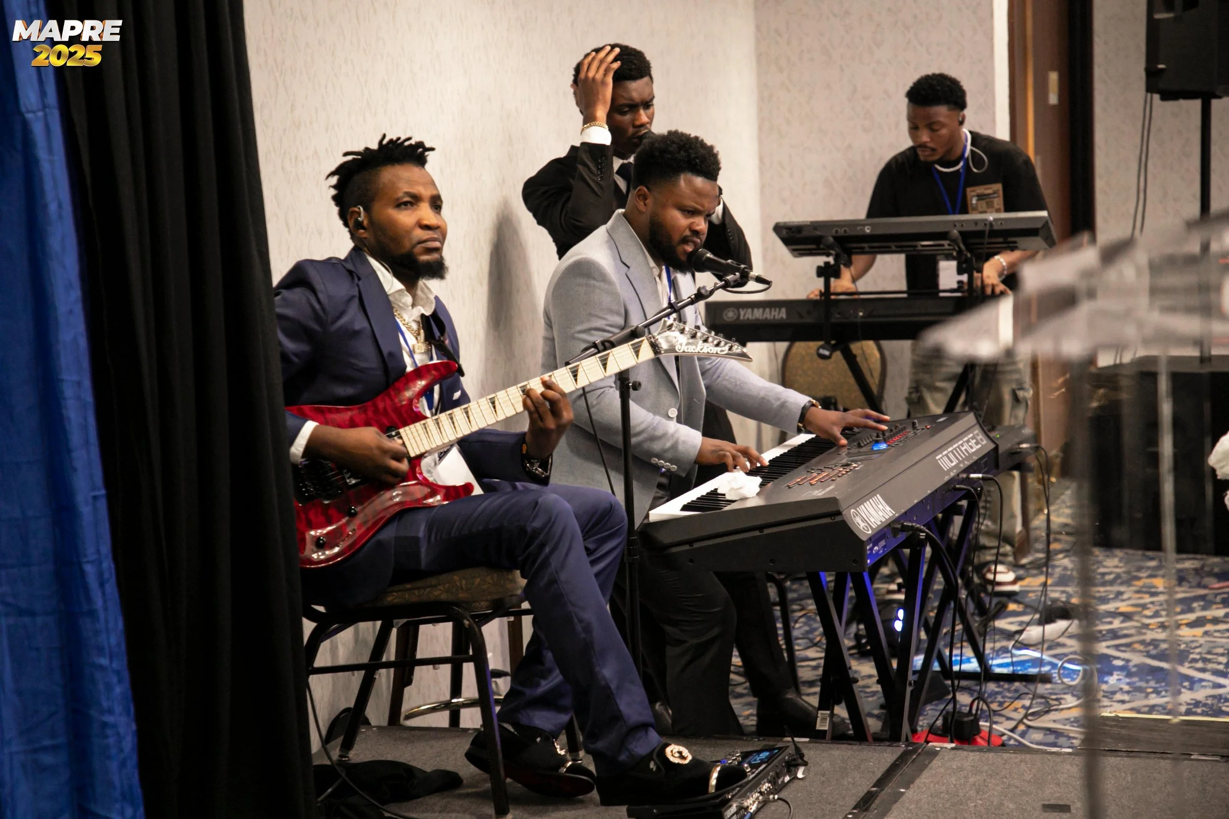 Four men performing music on a stage, one playing an electric guitar, one on a keyboard, and two standing, with one appearing to sing or speak into a microphone.