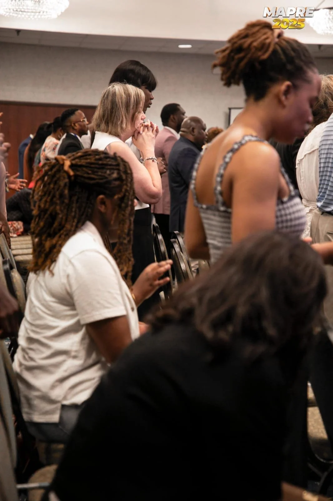 A diverse group of people attending a prayer or meditation session in a conference room, with some individuals standing, eyes closed, and hands clasped or in prayer.