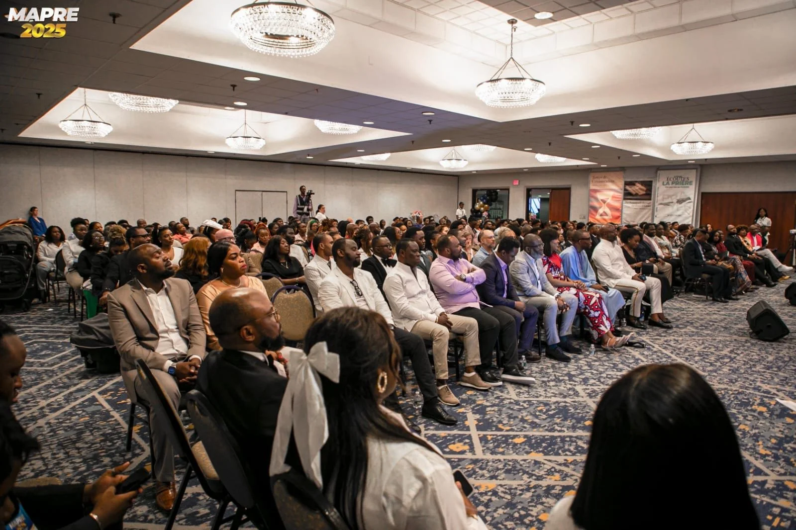 A large group of people seated in a conference room, attending a formal event or seminar, with some standing at the back and one person filming. The room is decorated with chandeliers and banners, including one that reads 'MAPRE 2025.'