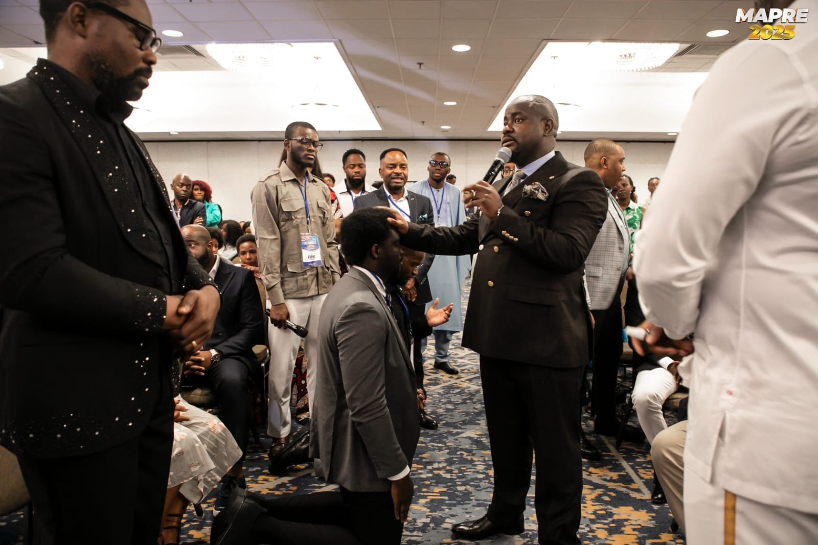 A group of people gathered at a conference, with one man in a black suit leading a prayer while kneeling, others standing around him with some placing hands on his head and shoulders, in a hotel event room.