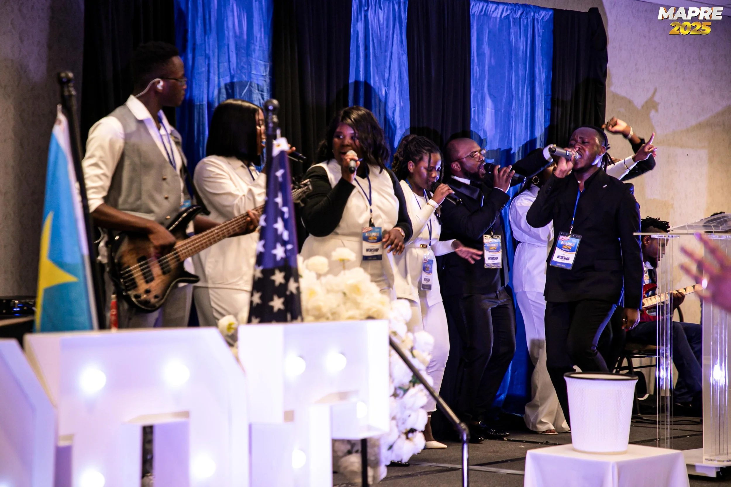 A group of seven people on stage, some singing and playing musical instruments, at a conference or event with a blue and black curtain backdrop, and a banner in the top right corner that says "MAPRE 2025."