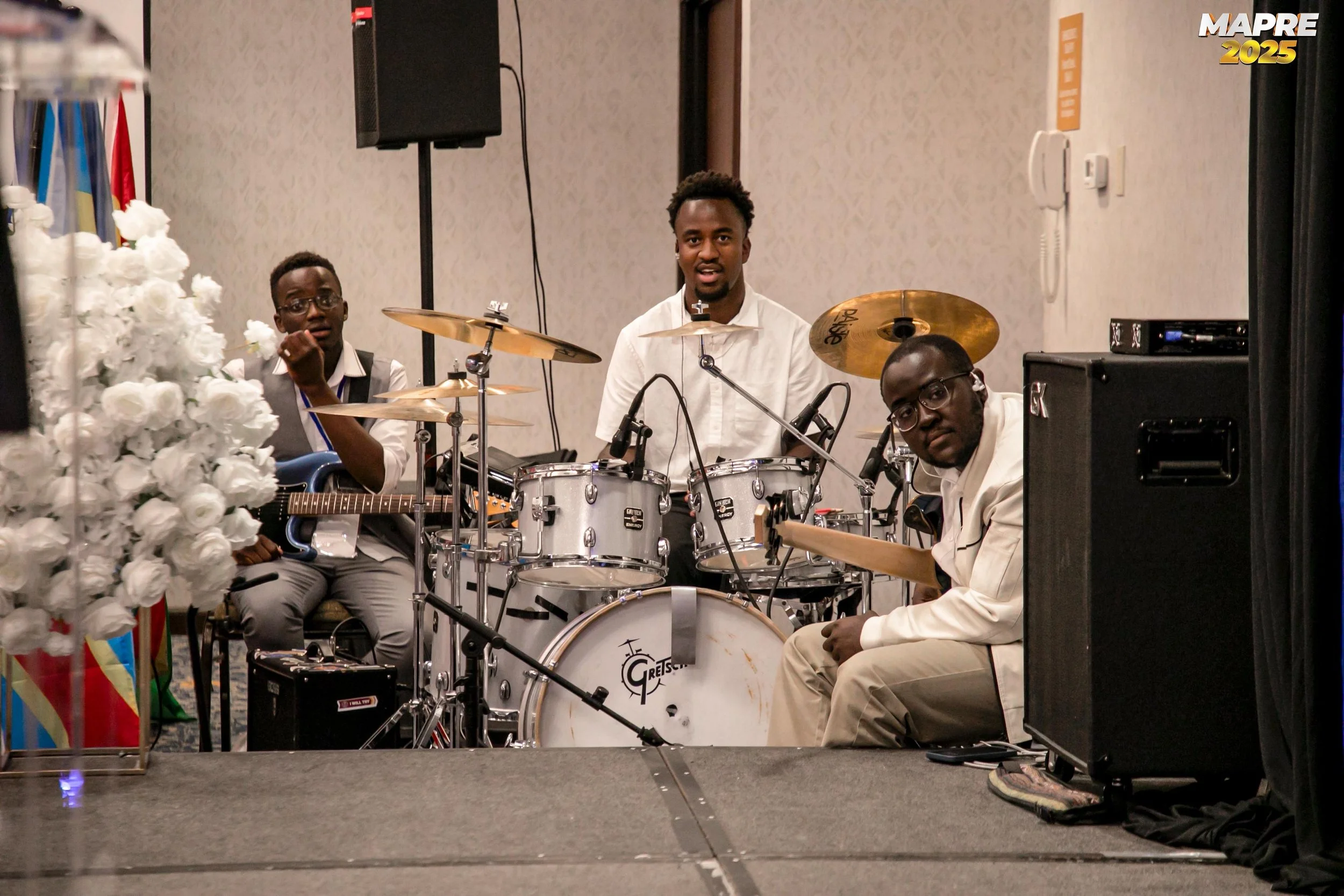 Three musicians performing at an indoor event. The man in the center is playing a drum set and wearing a white shirt. To his left, a young man with glasses is sitting with a guitar, and to his right, another man with glasses is sitting, possibly adjusting equipment. The background features a patterned wall, speakers, and a floral arrangement on the left. The top right corner has a logo that reads 'MAPRE 2025'.