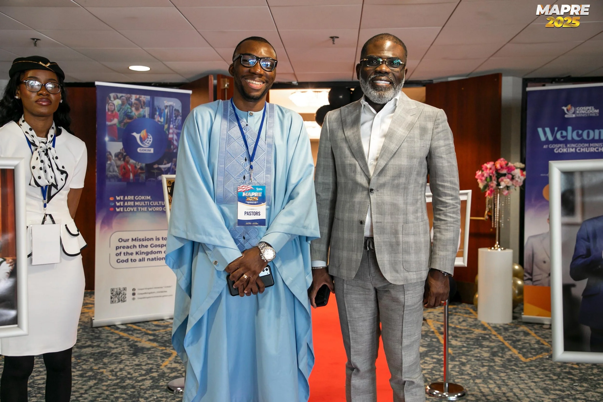 Three men and one woman standing at a conference or event, smiling for the camera. They are dressed in formal and traditional attire. Background displays banners with information about Gospel Kingdom Ministries Intl and a welcome message.
