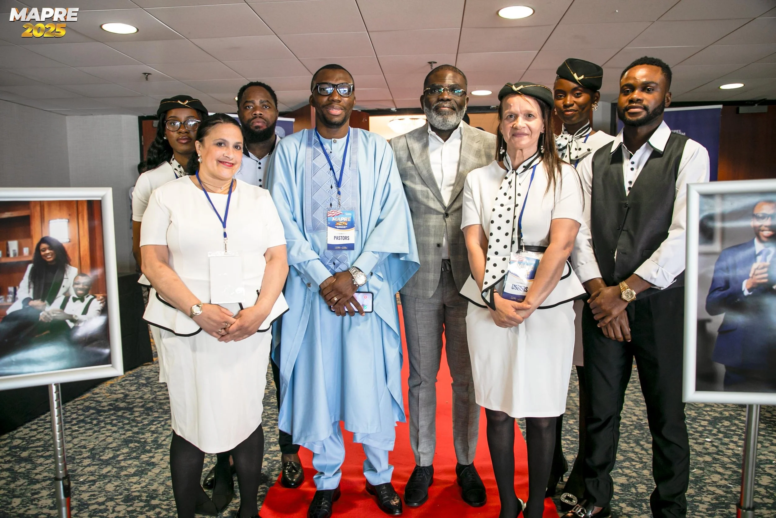 A diverse group of eight people standing on a red carpet at an event, with some wearing formal attire and others in coordinated outfits. Two people are wearing black hats with gold accents, and all are smiling or looking serious. There are framed photographs on stands beside them, and a banner in the background that reads 'MAPRE 2025'.