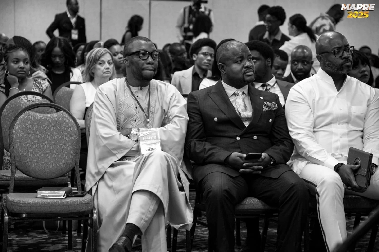 A diverse group of people attending a conference, seated in rows of chairs, with some wearing identification lanyards. The front row includes a man in traditional attire, a man in a suit, and a man in white, with some attendees focused or looking ahead.
