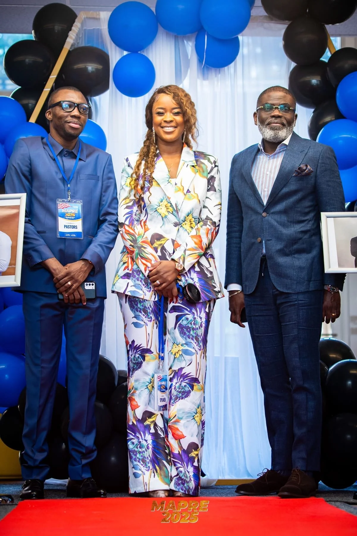 Three people standing in front of blue and black balloons, smiling for the camera at an event called MAPRE 2025. The man on the left wears a blue suit and glasses, the woman in the middle wears a colorful floral pantsuit, and the man on the right wears a dark blue suit and glasses. A red carpet with yellow text reading "MAPRE 2025" is visible at the bottom.