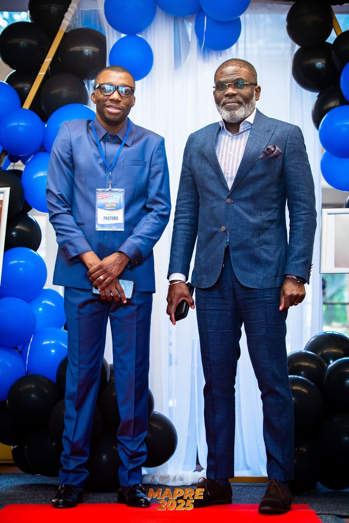 Two men in formal suits standing on a red carpet, surrounded by blue and black balloons, at an event called MAPRE 2025.