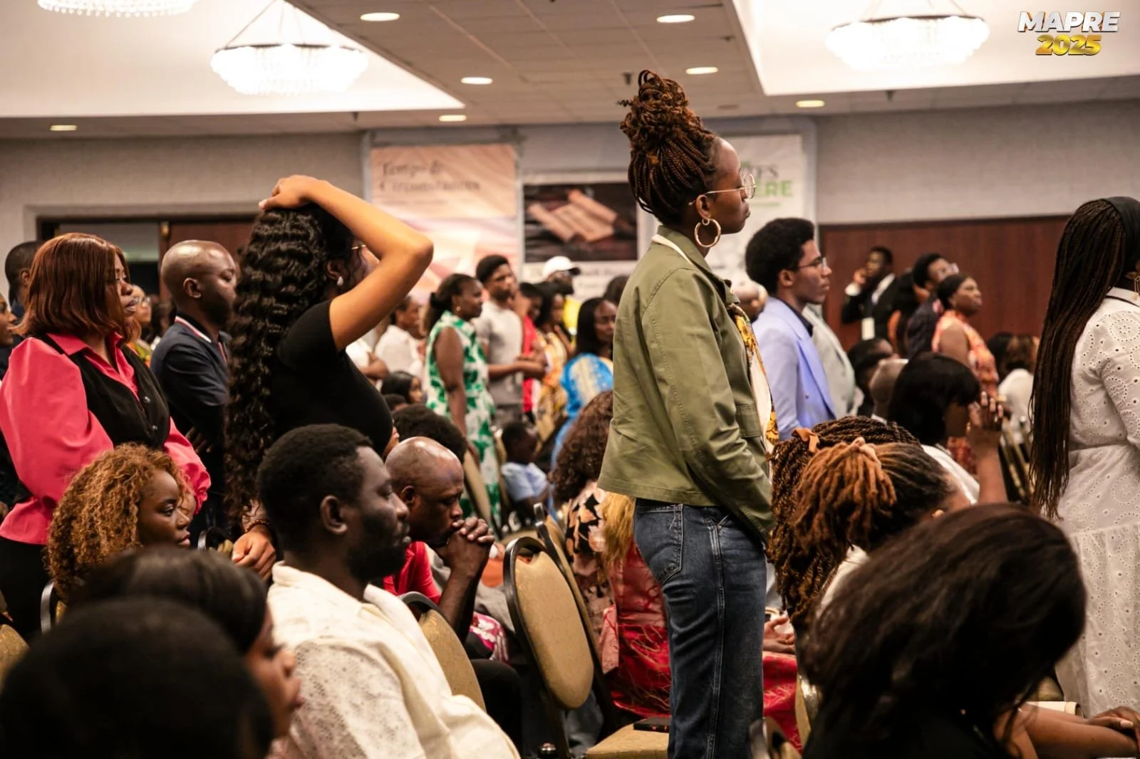 A large audience of men and women attending a conference or event in a ballroom, with many standing and some seated, focused on a speaker or presentation.