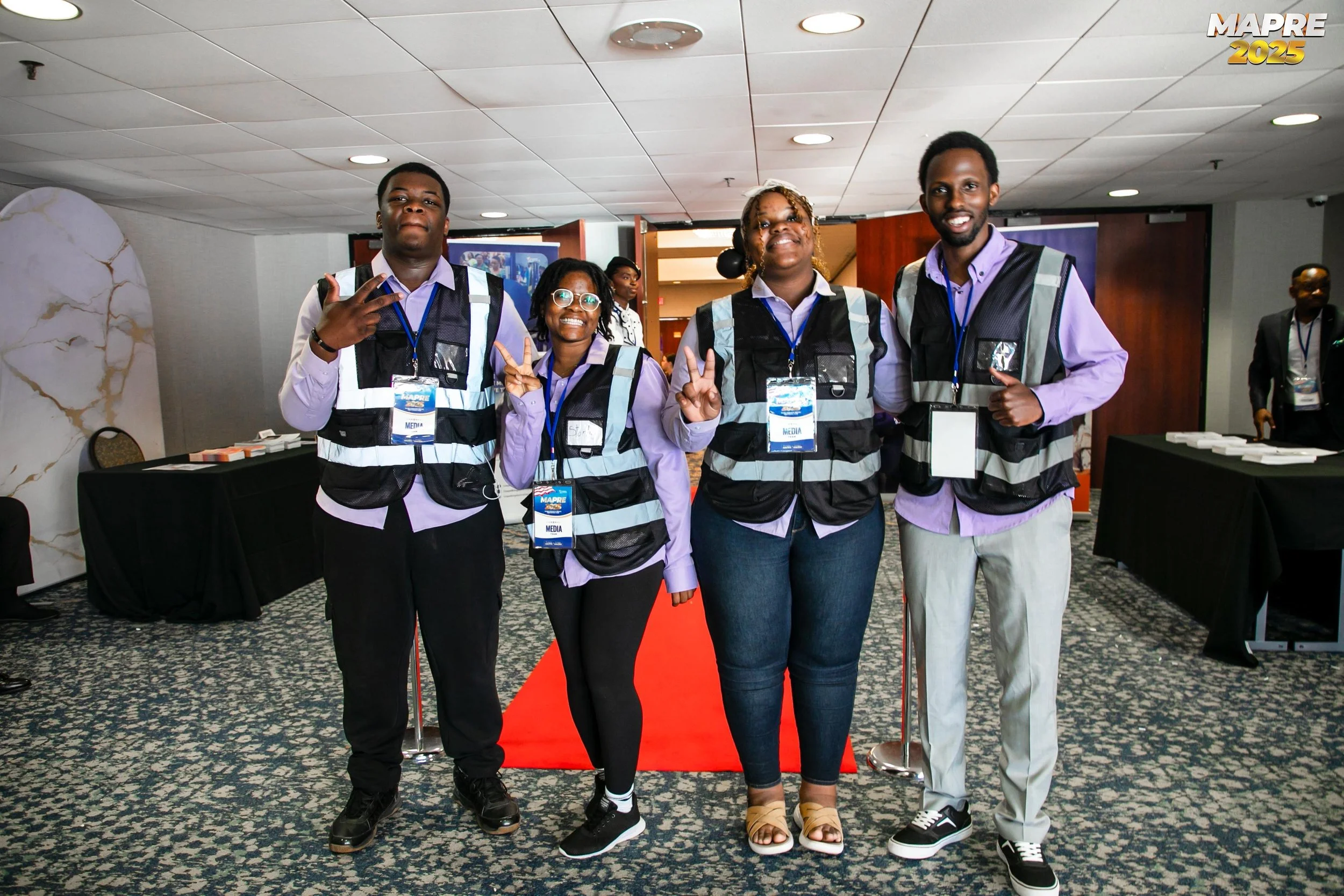 Four people standing on a red carpet at an indoor event, all wearing purple shirts and black vests with media badges, smiling and making peace signs or gestures, with a conference room setting in the background.