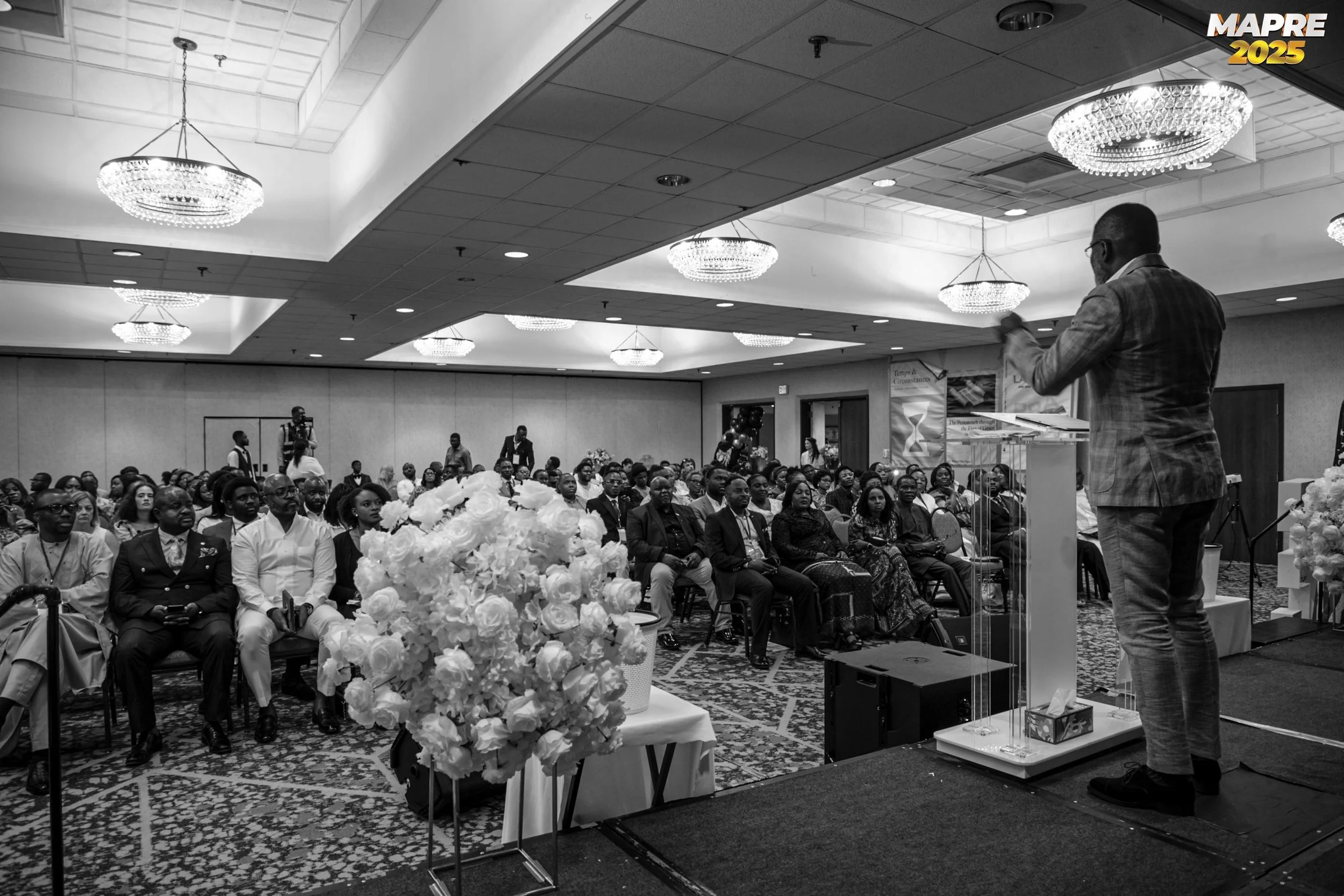 A speaker in a suit addressing an audience seated in a conference room with chandeliers and floral arrangements.
