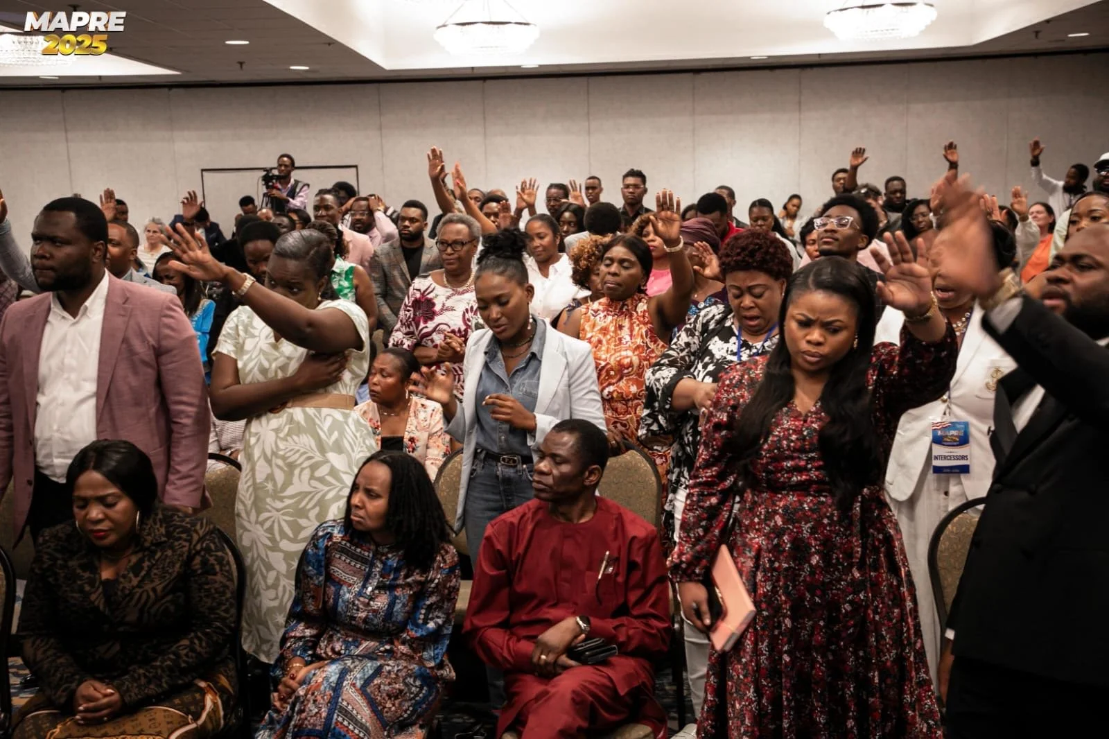 A large group of people, mostly African, gathered in a conference room, with many raising their hands and some with eyes closed, possibly during prayer or worship. Some are sitting while others are standing.